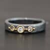 Photo of the "Adoration" Ring. Made out of sterling silver, gold, and diamonds. There are three diamonds that go straight across the center of the ring. Each one is set in gold, and each diamond is separated by a ball of gold. There are four balls of gold in total. The rest of the ring is oxidized and has a textured look to it.