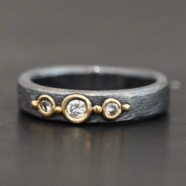 Photo of the "Adoration" Ring. Made out of sterling silver, gold, and diamonds. There are three diamonds that go straight across the center of the ring. Each one is set in gold, and each diamond is separated by a ball of gold. There are four balls of gold in total. The rest of the ring is oxidized and has a textured look to it.