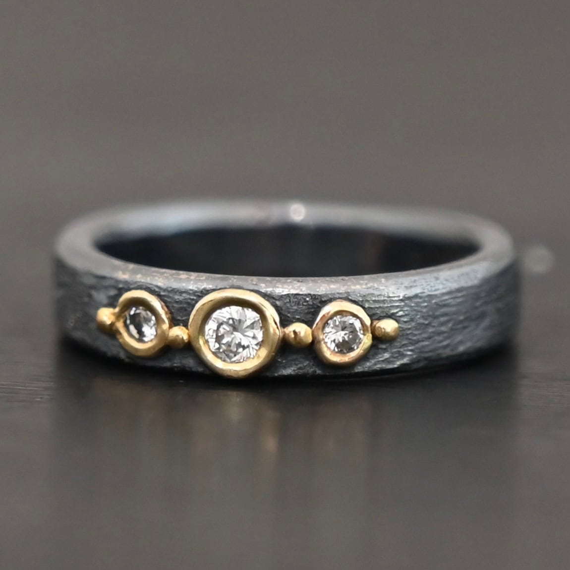 Photo of the "Adoration" Ring. Made out of sterling silver, gold, and diamonds. There are three diamonds that go straight across the center of the ring. Each one is set in gold, and each diamond is separated by a ball of gold. There are four balls of gold in total. The rest of the ring is oxidized and has a textured look to it.