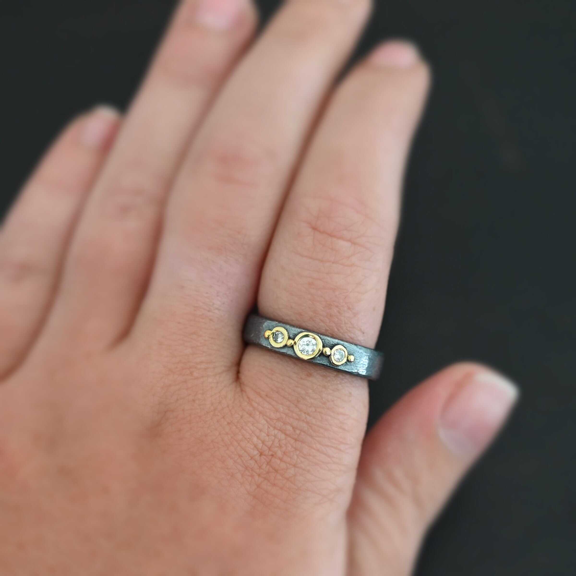Photo of the "Adoration" Ring. Made out of sterling silver, gold, and diamonds. There are three diamonds that go straight across the center of the ring. Each one is set in gold, and each diamond is separated by a ball of gold. There are four balls of gold in total. The rest of the ring is oxidized and has a textured look to it. Ring is shown up close worn by a model.