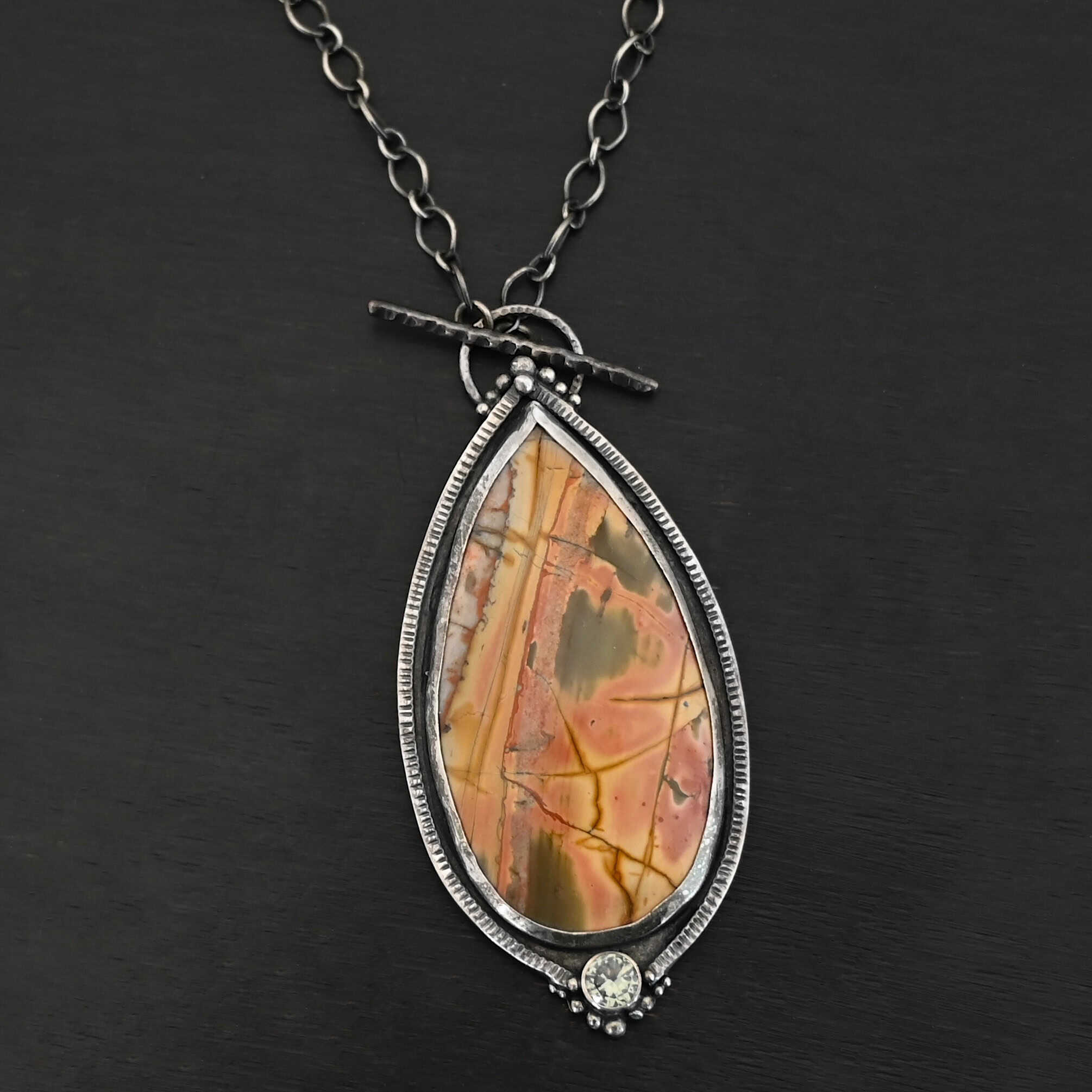 Finding You Necklace "Finding You" Necklace. There is a large, pear shaped cherry creek jasper, pointing upwards. There is a small circular green topaz beneath it, and silver ball detail around that. The silver around the rest of the stone is hammered with lines. There is silver ball detail on the top as well. Necklace is a toggle clasp.