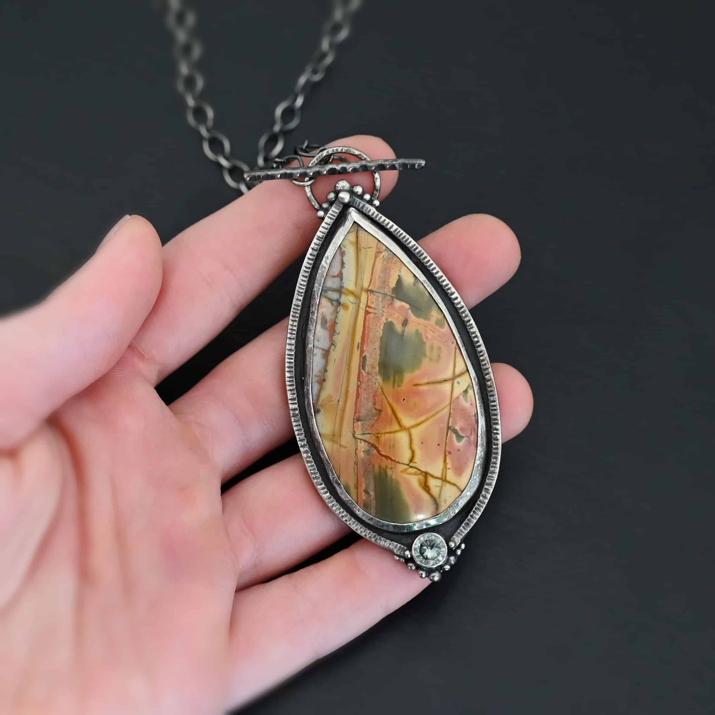 Finding You Necklace "Finding You" Necklace. There is a large, pear shaped cherry creek jasper, pointing upwards. There is a small circular green topaz beneath it, and silver ball detail around that. The silver around the rest of the stone is hammered with lines. There is silver ball detail on the top as well. Necklace is a toggle clasp.