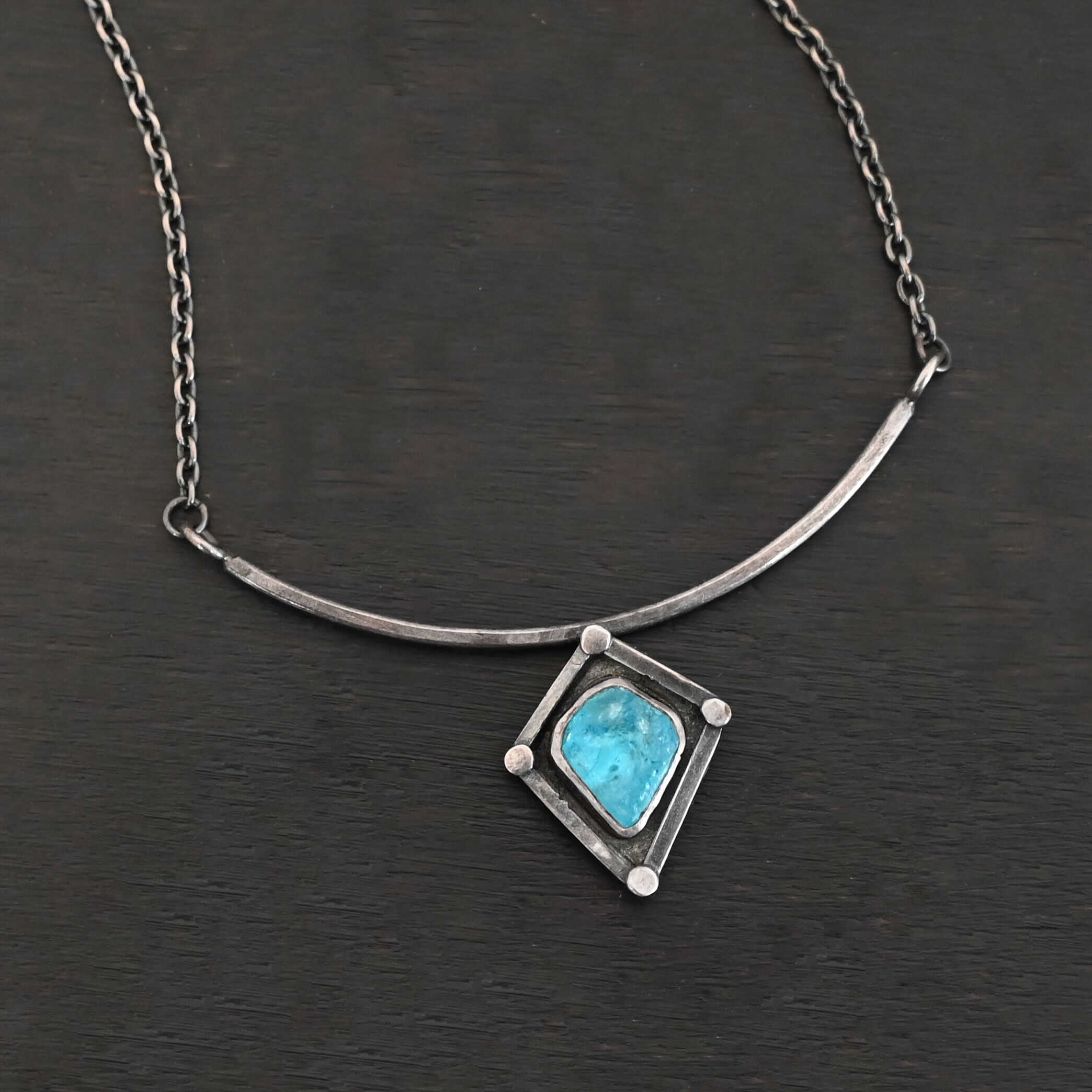 Elsa Necklace "Elsa" Necklace. There is a rough cut apatite in the center of a diamond shaped piece of silver. There is a dot of silver on each point of the silver diamond. The silver diamond is soldered to a curved bar of silver.