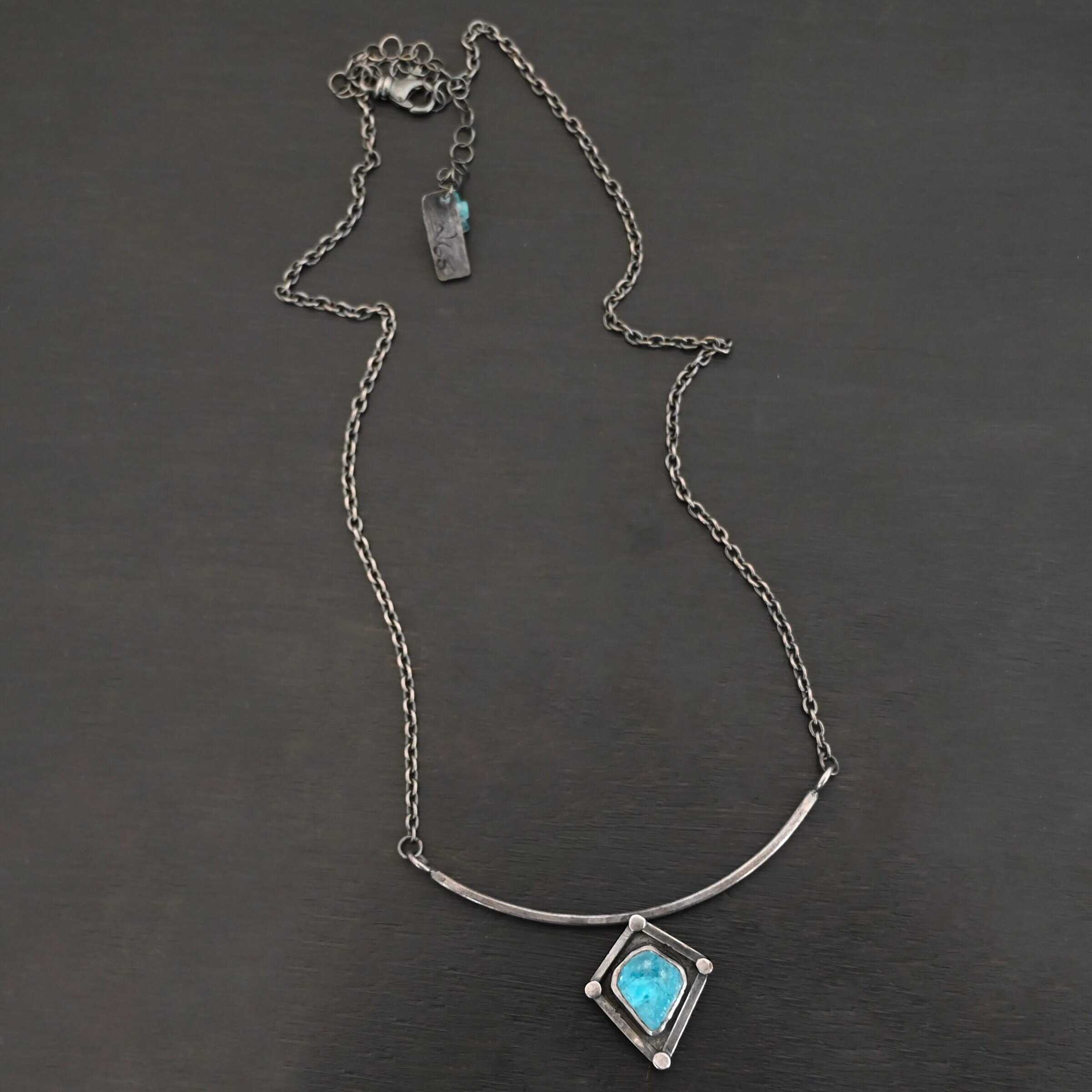 Elsa Necklace "Elsa" Necklace. There is a rough cut apatite in the center of a diamond shaped piece of silver. There is a dot of silver on each point of the silver diamond. The silver diamond is soldered to a curved bar of silver.