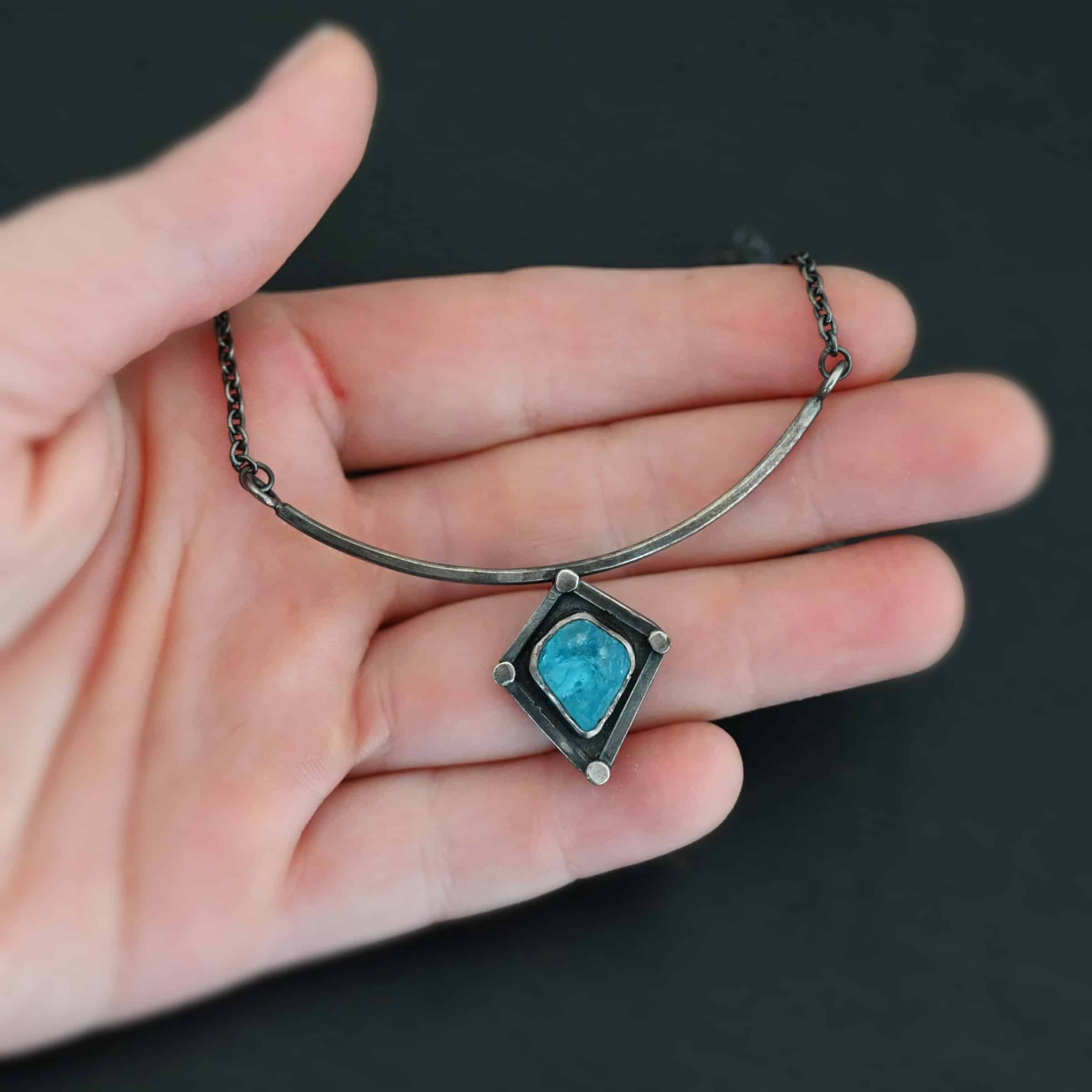 Elsa Necklace "Elsa" Necklace. There is a rough cut apatite in the center of a diamond shaped piece of silver. There is a dot of silver on each point of the silver diamond. The silver diamond is soldered to a curved bar of silver.