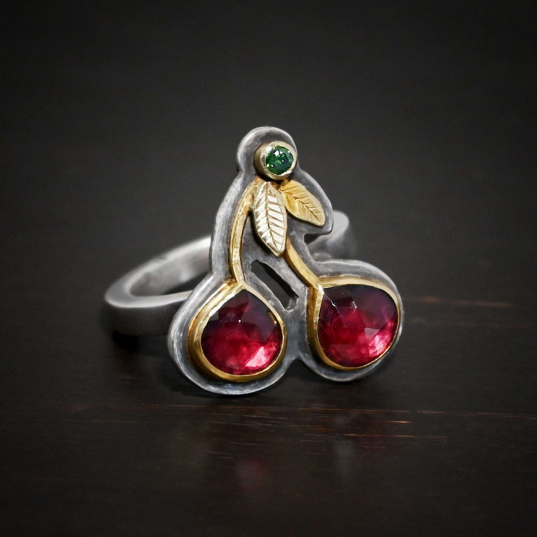 Door County Cherry Ring Close up of the "Door County Cherry" Ring. Made out of sterling silver, gold, garnets, and green diamond. There are two slightly teardrop shaped garnets that make up the "Cherry" part of the ring. They are set in gold, and the gold leads up to the small circular green diamond as a part of the "stem" of the cherries. There are two gold leaves on the ring as well. Ring is set against a dark background.