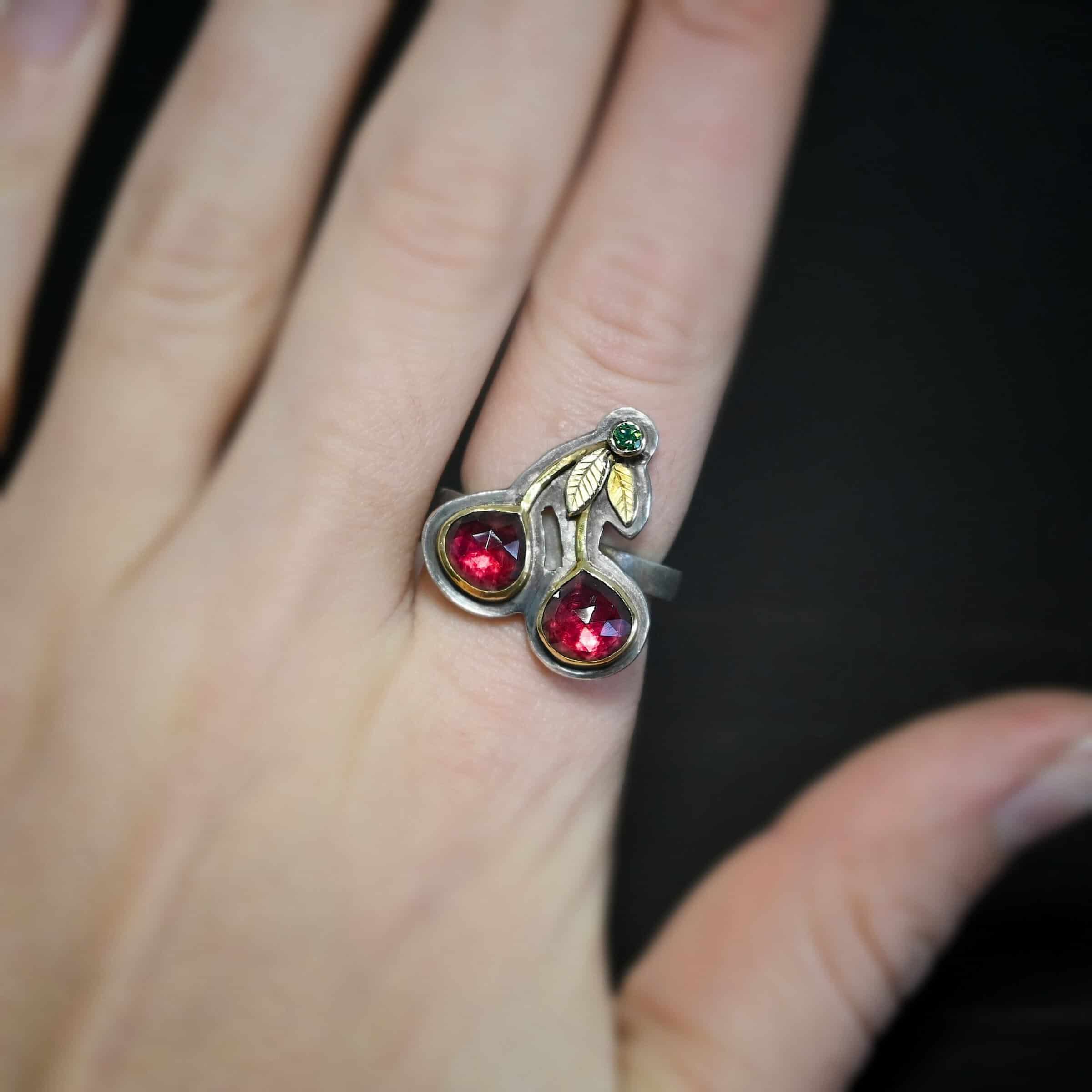 Door County Cherry Ring Close up of the "Door County Cherry" Ring. Made out of sterling silver, gold, garnets, and green diamond. There are two slightly teardrop shaped garnets that make up the "Cherry" part of the ring. They are set in gold, and the gold leads up to the small circular green diamond as a part of the "stem" of the cherries. There are two gold leaves on the ring as well. Ring is worn by a model on their pointer finger to demonstrate the size of the ring.