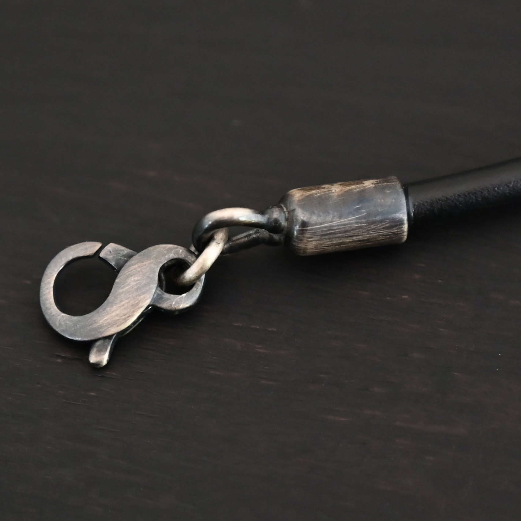 Clasp for the "Wren Wire" Chain. Clasp is made out of oxidized sterling silver and is attached to oxidized sterling silver end cap. Clasp is a figure-eight shape, and shown against a dark background.