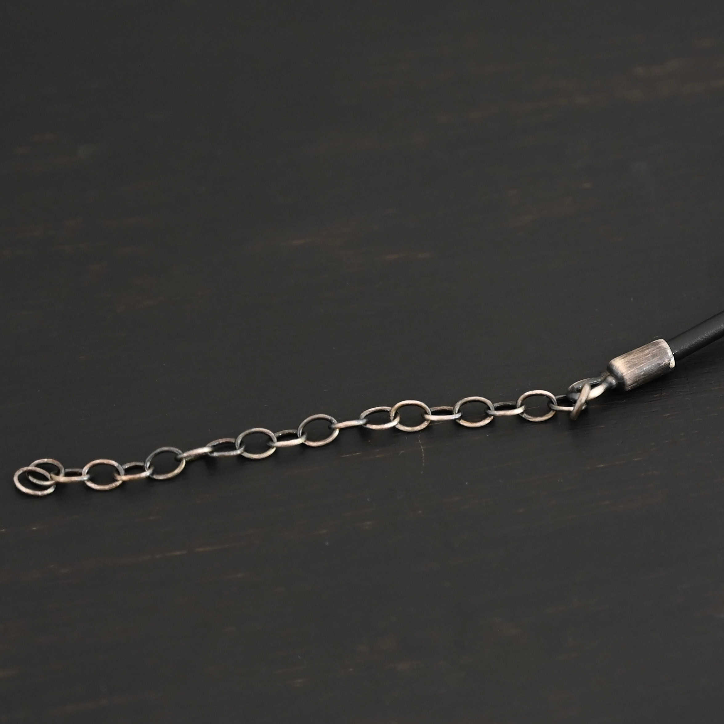 "Wren Wire" Chain extension. Extension is shown so you can make the chain longer or shorter. Extension is made out of solid sterling silver and is oxidized. It is attached to a sterling silver end cap that is also oxidized. Extension is shown against a dark background.