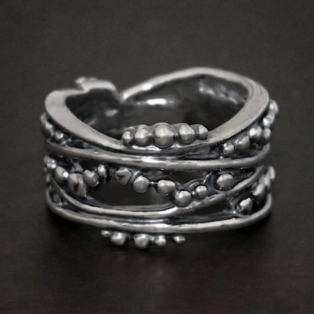 River Ring Photo of the "River" Ring. Made out of sterling silver. It has multiple lines of silver that intercept at random places, but all of them meet at the back to create the band of the ring. There are silver dots scattered all over the ring in between the gaps of where the silver does not meet. The top and bottom of the lines flow and create an uneven ring band. Ring is sitting against a dark background.