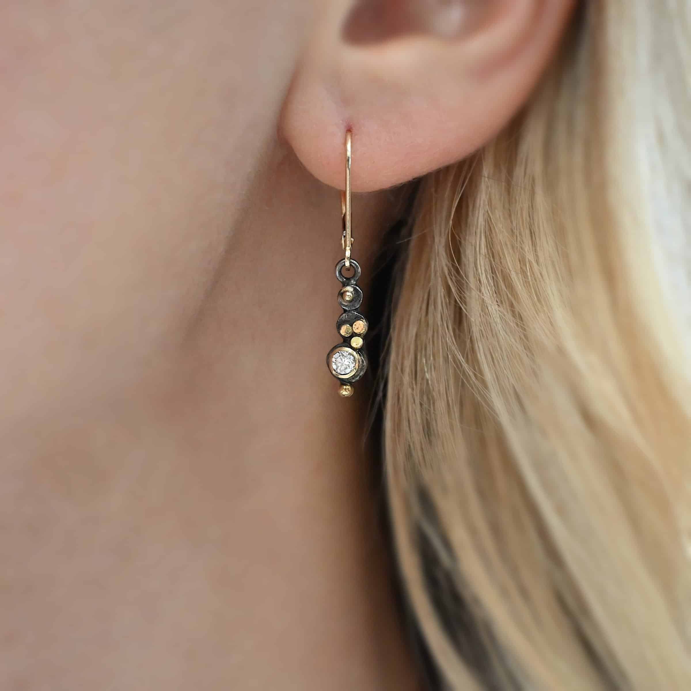 Prairie Princess Earrings Close up of the "Prairie Princess" Earrings. Made out of sterling silver, gold, and diamonds. One earring is shown on a model. There are three silver dots that are flat, going from smallest to biggest from the top to the bottom. On the smallest top silver hammered dot is a gold dot that is not hammered. On the second hammered dot are three gold dots that are hammered. On the final hammered dot is a diamond that is set in gold. On the bottom of the earring is a gold dot. Earrings are on gold leverback ear wires. Model is wearing the earring to demonstrate length.