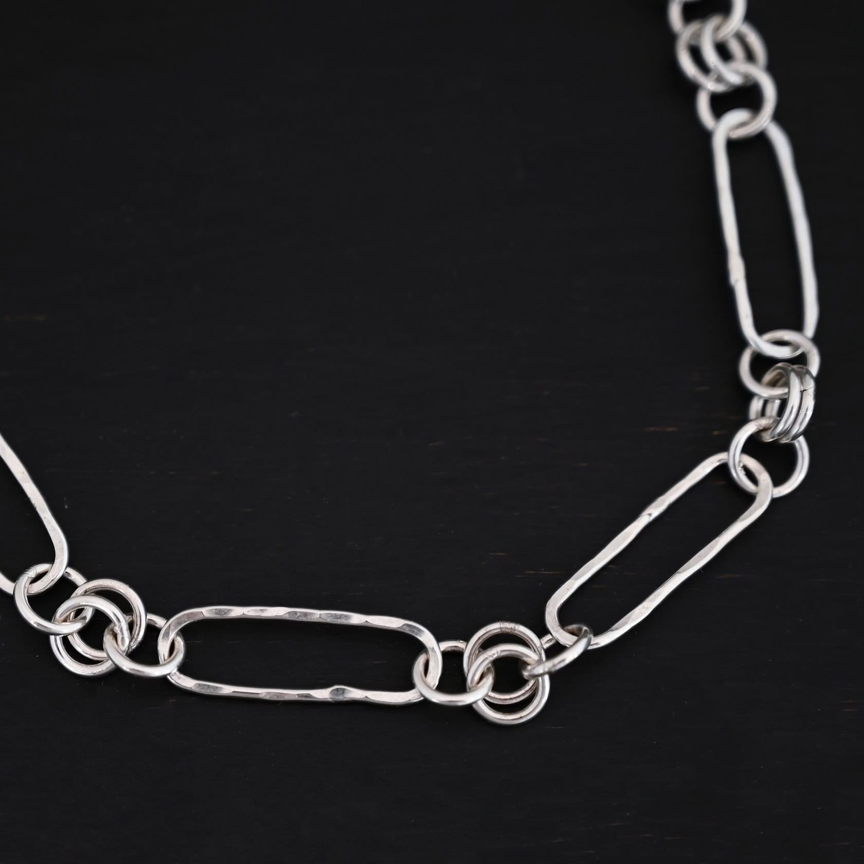 Close up of the "Bekah" Chain This chain is handmade out of sterling silver and is shiny. This chain has large oval links that are hammered and connected together by four jump rings. The chain is laying on a dark background.