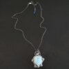 Photo of the "Lake Monona" Necklace. Made out of sterling silver, leland blue, and aquamarine. The leland blue is the center stone. It is a large teardrop shape with the tip of the teardrop pointing downwards. It is set in setting silver. There are groups of three silver balls scattered all around the rest of the silver setting. There are five groups of silver balls, and each group has three balls. There is a small circular aquamarine set in silver on the top right of the necklace on the silver setting. On the back of the necklace is a loop. There are three jump rings attached to the loop, and the chain of the necklace goes through them. Necklace is laying against a dark background.