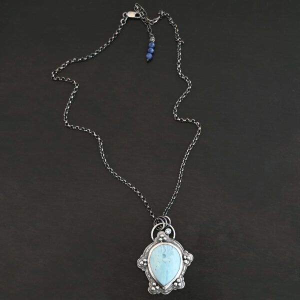 Photo of the "Lake Monona" Necklace. Made out of sterling silver, leland blue, and aquamarine. The leland blue is the center stone. It is a large teardrop shape with the tip of the teardrop pointing downwards. It is set in setting silver. There are groups of three silver balls scattered all around the rest of the silver setting. There are five groups of silver balls, and each group has three balls. There is a small circular aquamarine set in silver on the top right of the necklace on the silver setting. On the back of the necklace is a loop. There are three jump rings attached to the loop, and the chain of the necklace goes through them. Necklace is laying against a dark background.
