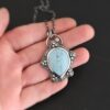 Photo of the "Lake Monona" Necklace. Made out of sterling silver, leland blue, and aquamarine. The leland blue is the center stone. It is a large teardrop shape with the tip of the teardrop pointing downwards. It is set in setting silver. There are groups of three silver balls scattered all around the rest of the silver setting. There are five groups of silver balls, and each group has three balls. There is a small circular aquamarine set in silver on the top right of the necklace on the silver setting. On the back of the necklace is a loop. There are three jump rings attached to the loop, and the chain of the necklace goes through them. Necklace is laying in the fingertips of a hand to demonstrate size.