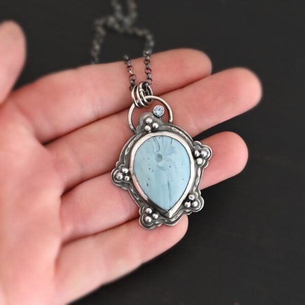 Photo of the "Lake Monona" Necklace. Made out of sterling silver, leland blue, and aquamarine. The leland blue is the center stone. It is a large teardrop shape with the tip of the teardrop pointing downwards. It is set in setting silver. There are groups of three silver balls scattered all around the rest of the silver setting. There are five groups of silver balls, and each group has three balls. There is a small circular aquamarine set in silver on the top right of the necklace on the silver setting. On the back of the necklace is a loop. There are three jump rings attached to the loop, and the chain of the necklace goes through them. Necklace is laying in the fingertips of a hand to demonstrate size.