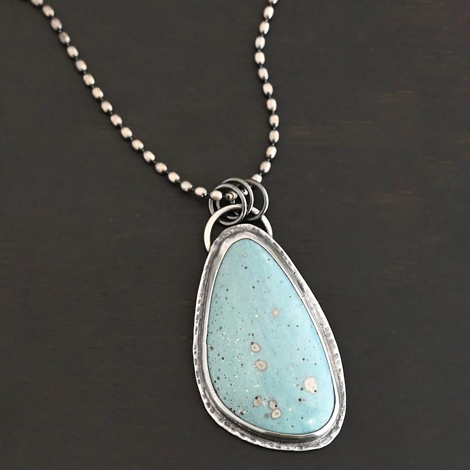 Close up of the "Galia" Necklace. Made out of sterling silver and leland blue. The leland blue is a light blue oval shape that is set vertically. There are some light gray dots scattered throughout the stone. There is a loop on the back of the pendant in which there are jump rings attached to it that are also attached to the chain of the necklace. The pendant is laying against a dark background.