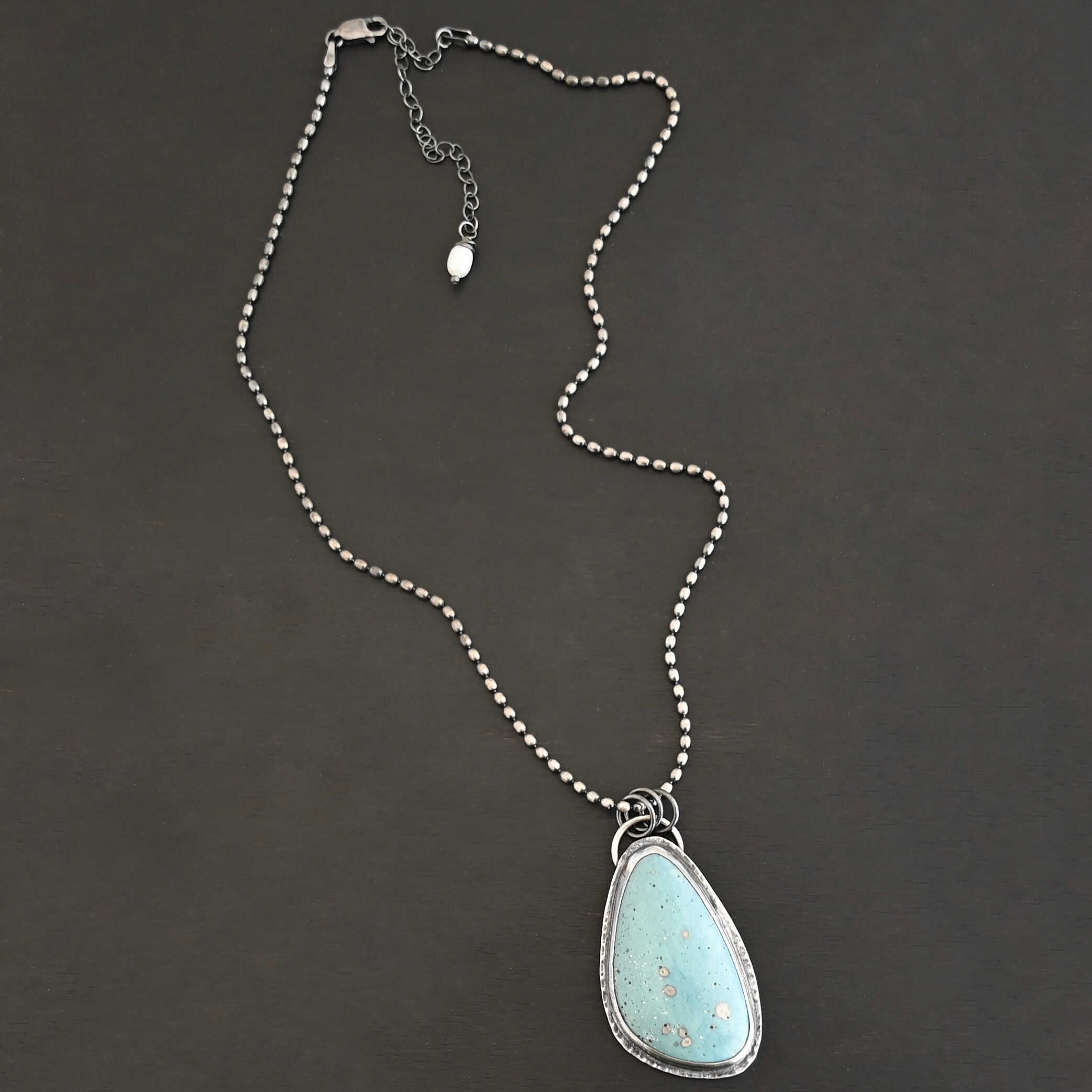 Close up of the "Galia" Necklace. Made out of sterling silver and leland blue. The leland blue is a light blue oval shape that is set vertically. There are some light gray dots scattered throughout the stone. There is a loop on the back of the pendant in which there are jump rings attached to it that are also attached to the chain of the necklace. The pendant is laying against a dark background.