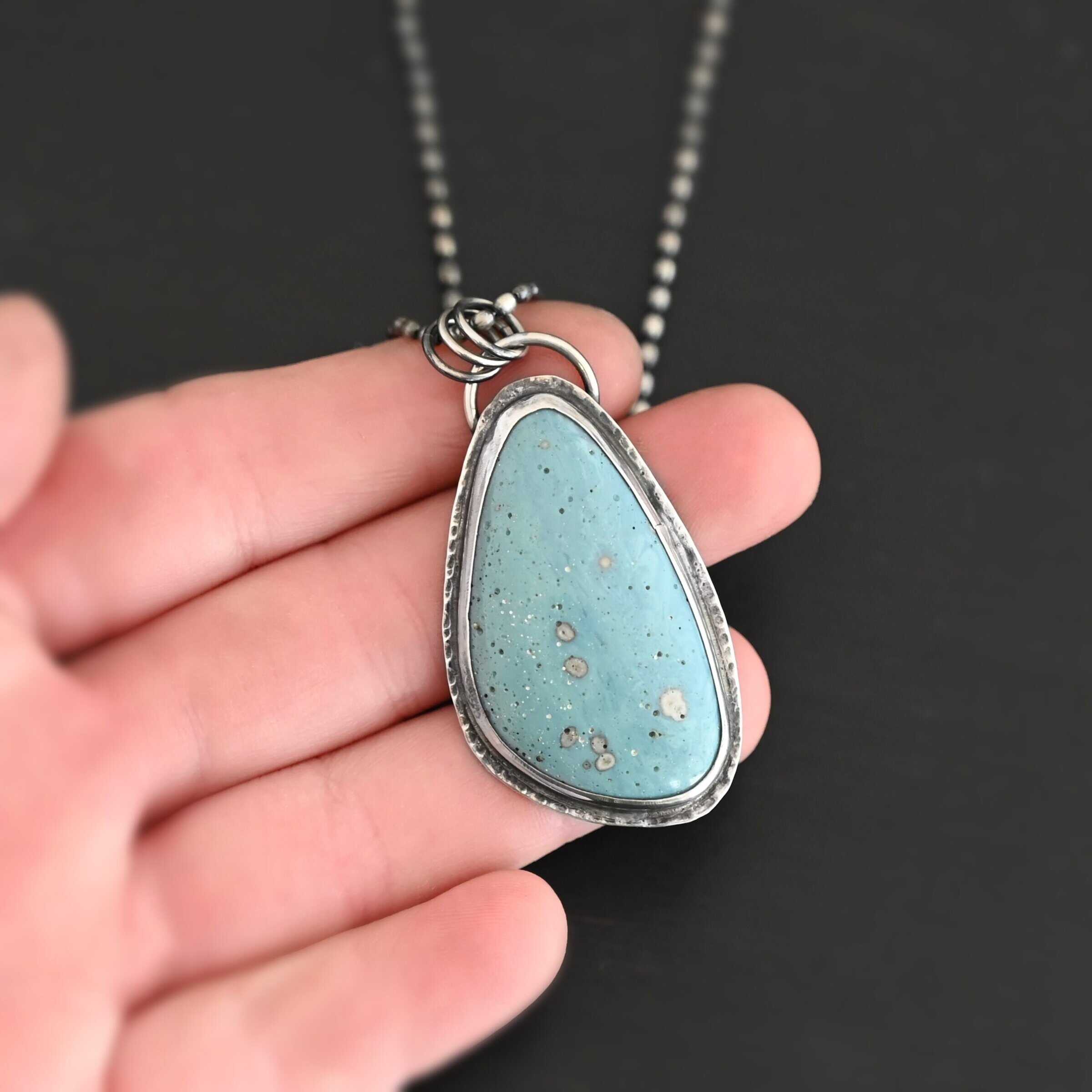 Close up of the "Galia" Necklace. Made out of sterling silver and leland blue. The leland blue is a light blue oval shape that is set vertically. There are some light gray dots scattered throughout the stone. There is a loop on the back of the pendant in which there are jump rings attached to it that are also attached to the chain of the necklace. Pendant is laying on the fingertips of a hand to demonstrate size.