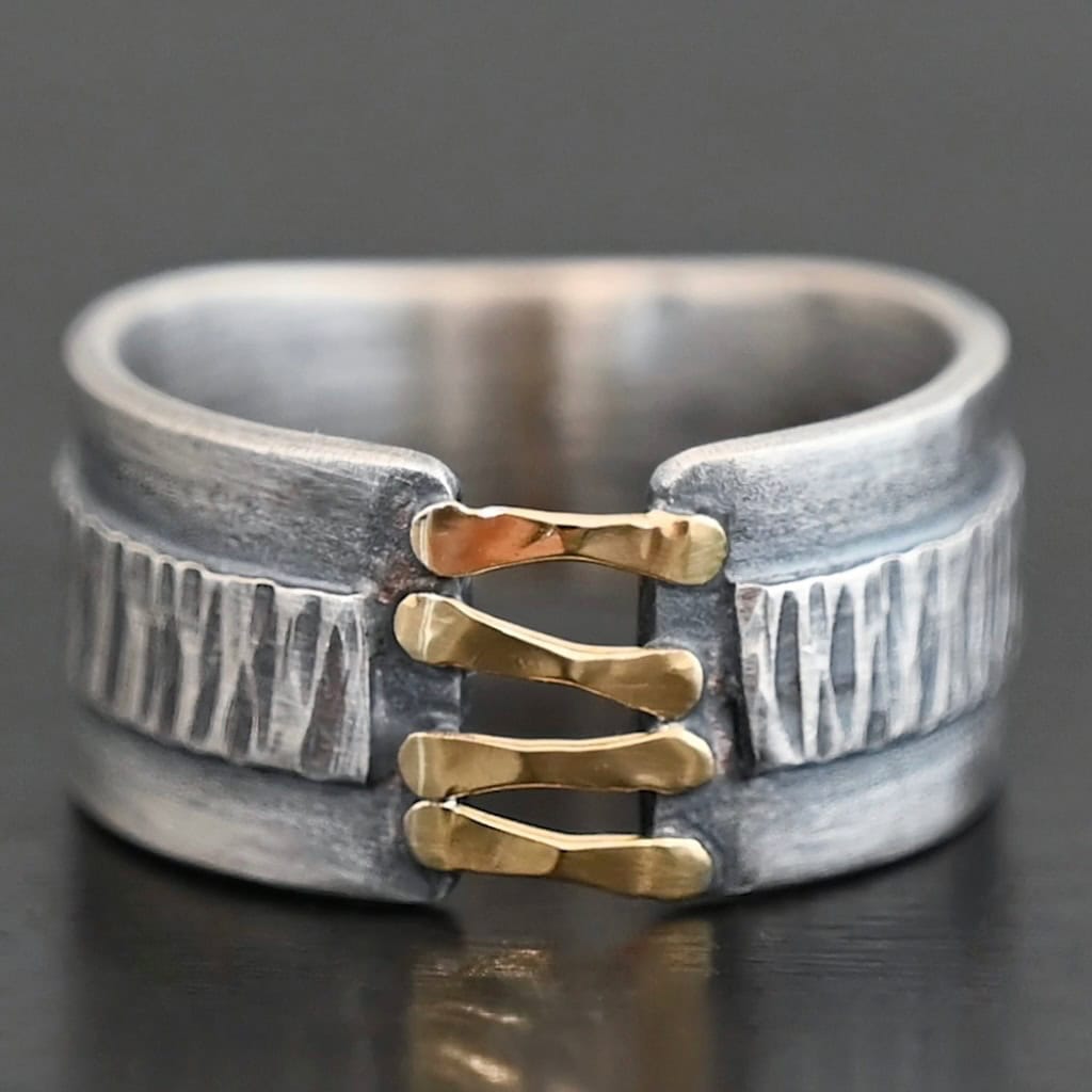 Photo of the "Boot Straps" Ring. Made out of sterling silver and gold. The band does not wrap all the way around. Connecting the two sides of the band are four hammered lines of gold. There is a thick rectangle of silver that wraps around the center of the band. It is engraved with wavy lines. Ring is sitting against a dark background.