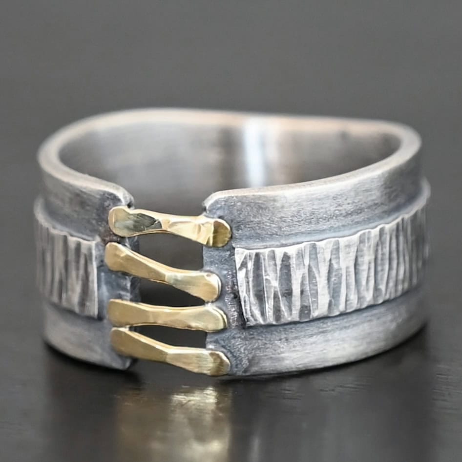 Photo of the "Boot Straps" Ring. Made out of sterling silver and gold. The band does not wrap all the way around. Connecting the two sides of the band are four hammered lines of gold. There is a thick rectangle of silver that wraps around the center of the band. It is engraved with wavy lines. Ring is sitting against a dark background.