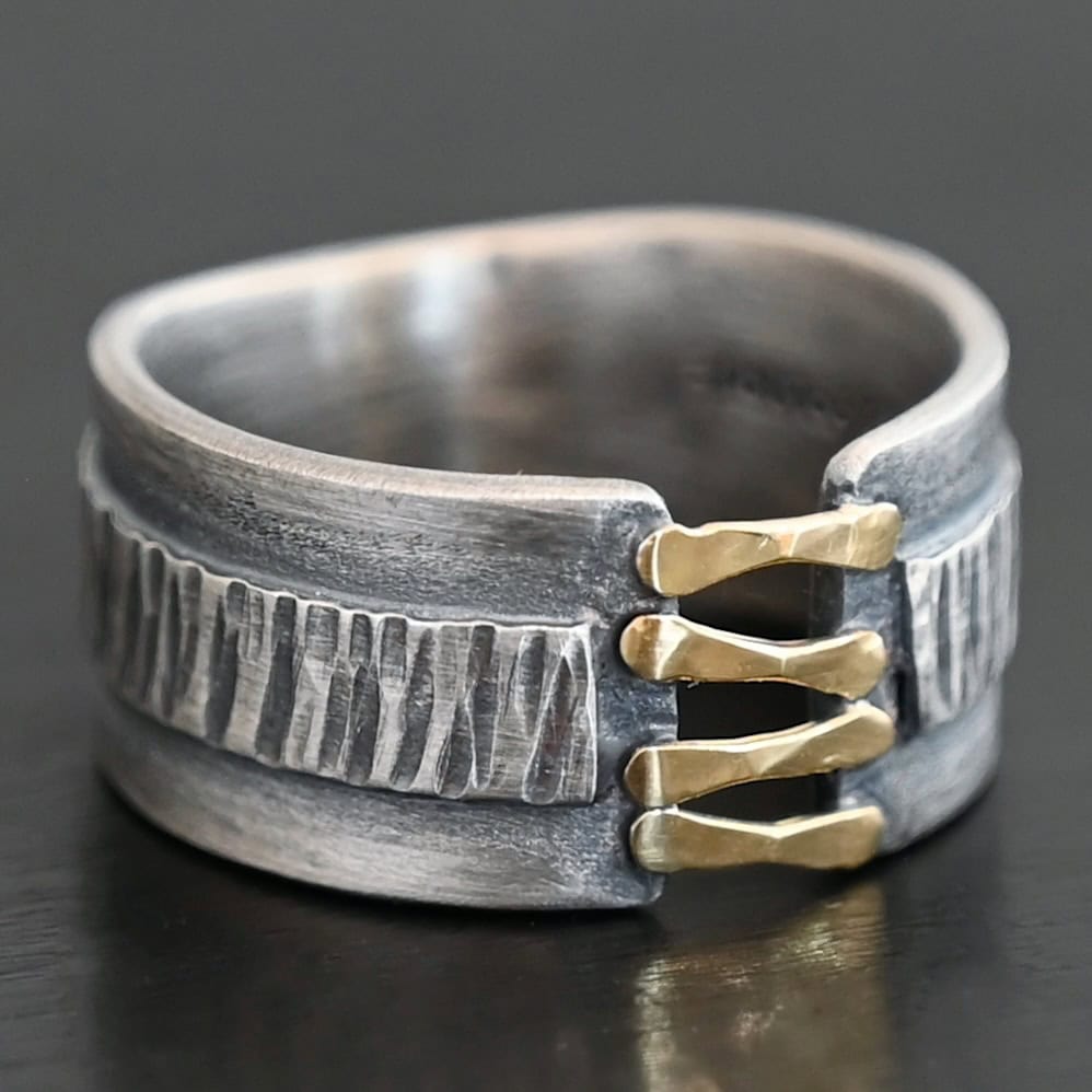 Photo of the "Boot Straps" Ring. Made out of sterling silver and gold. The band does not wrap all the way around. Connecting the two sides of the band are four hammered lines of gold. There is a thick rectangle of silver that wraps around the center of the band. It is engraved with wavy lines. Ring is sitting against a dark background.