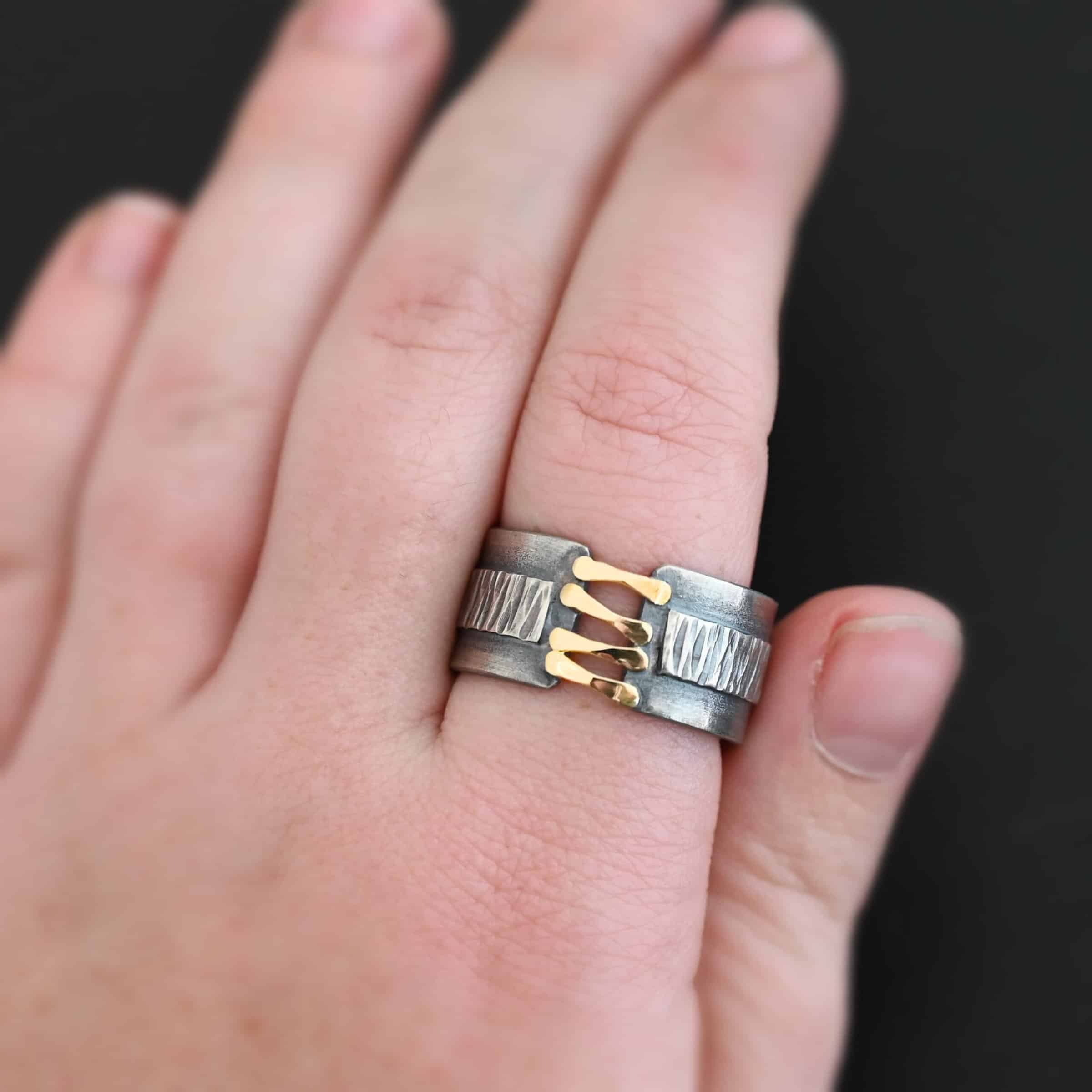 Photo of the "Boot Straps" Ring. Made out of sterling silver and gold. The band does not wrap all the way around. Connecting the two sides of the band are four hammered lines of gold. There is a thick rectangle of silver that wraps around the center of the band. It is engraved with wavy lines. Ring is worn by a model up close.