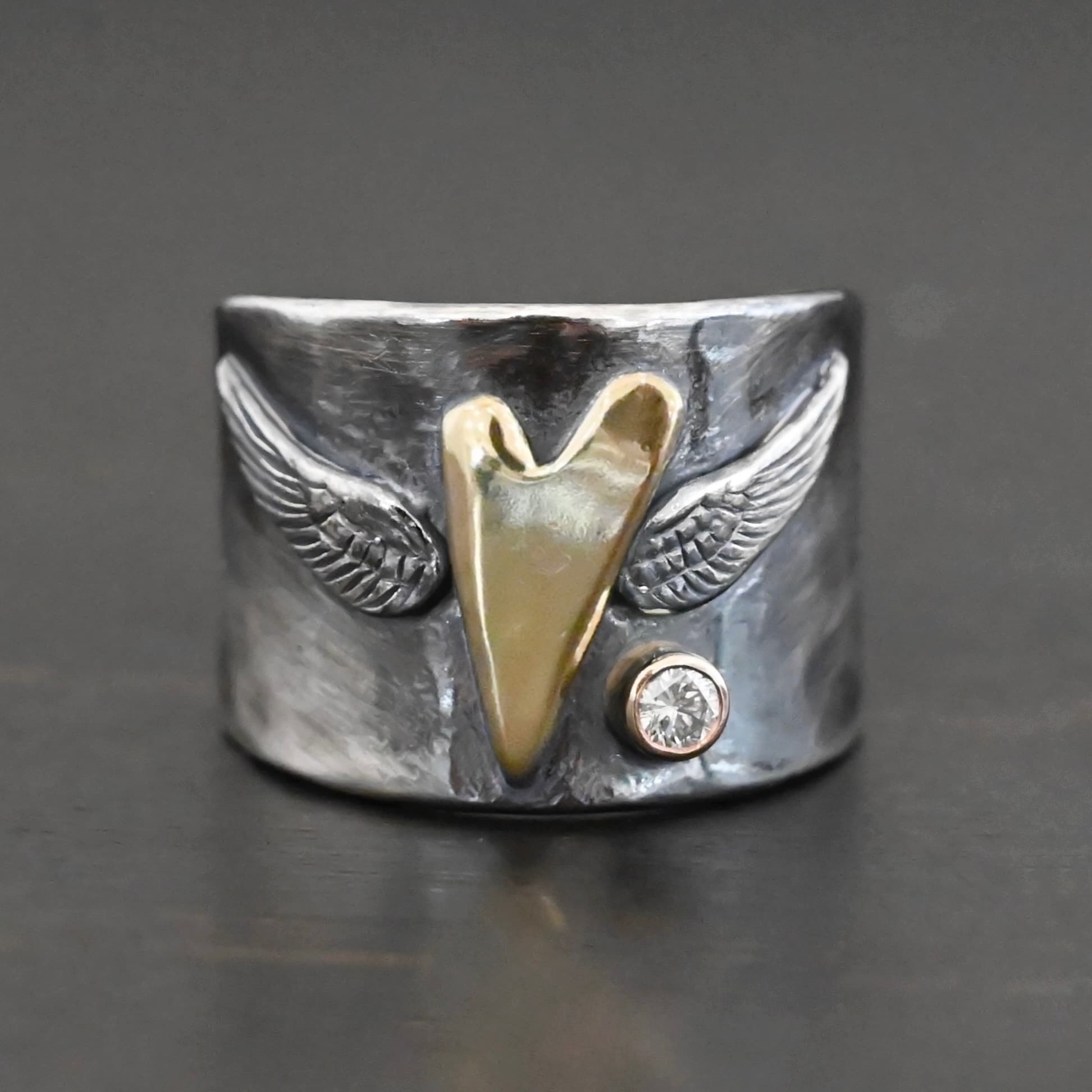 Close up of the "Wild Prairie" Ring. Made out of sterling silver, gold, and diamond. There is a gold heart in the center of the ring. It is a tall heart that stretches from almost the top of the ring to the bottom. There are sterling silver angel wings sprouting from each side of the heart. There is a diamond set in gold on the right side of the ring underneath one of the angel wings.
