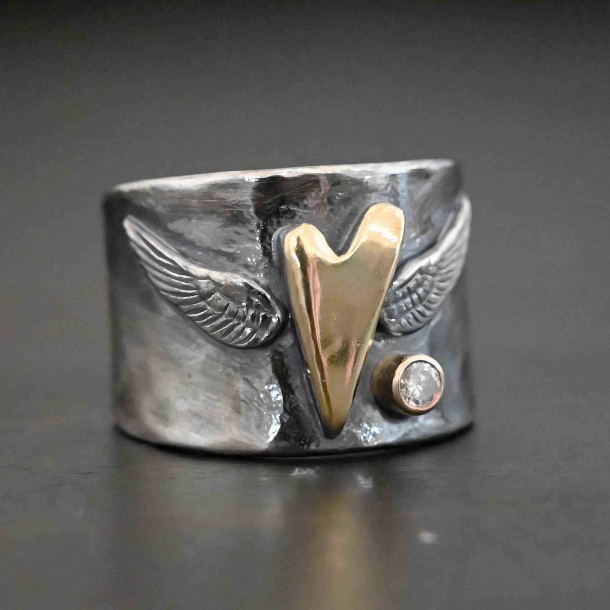Close up of the "Wild Prairie" Ring. Made out of sterling silver, gold, and diamond. There is a gold heart in the center of the ring. It is a tall heart that stretches from almost the top of the ring to the bottom. There are sterling silver angel wings sprouting from each side of the heart. There is a diamond set in gold on the right side of the ring underneath one of the angel wings.