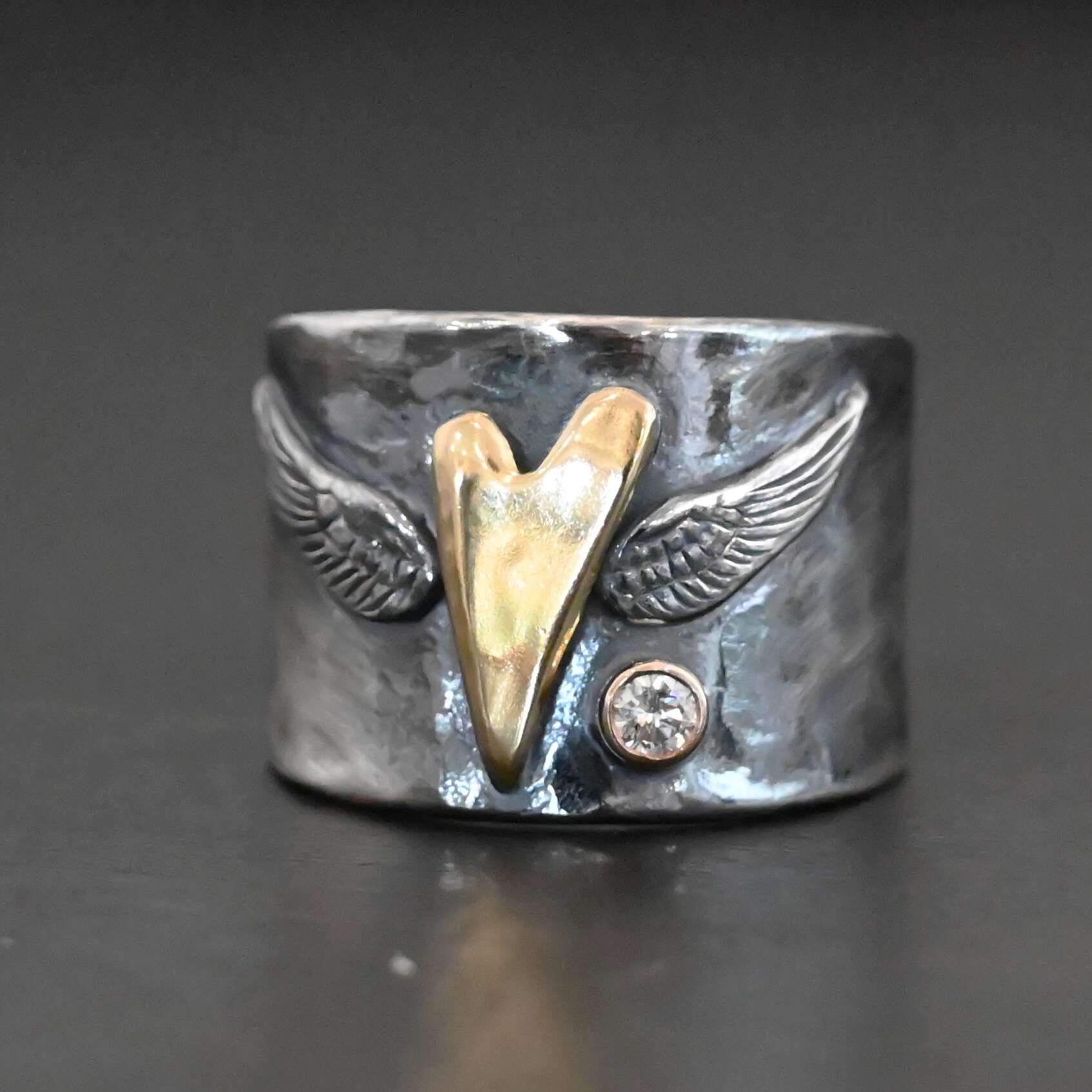 Close up of the "Wild Prairie" Ring. Made out of sterling silver, gold, and diamond. There is a gold heart in the center of the ring. It is a tall heart that stretches from almost the top of the ring to the bottom. There are sterling silver angel wings sprouting from each side of the heart. There is a diamond set in gold on the right side of the ring underneath one of the angel wings.