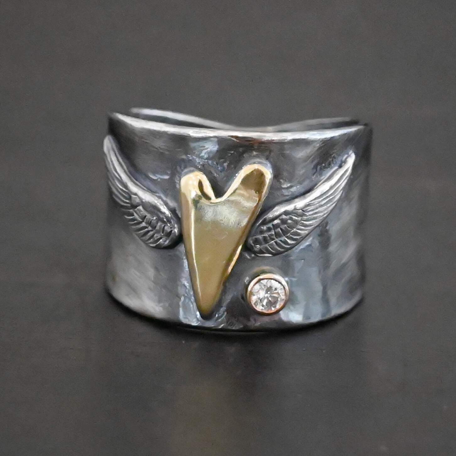 Close up of the "Wild Prairie" Ring. Made out of sterling silver, gold, and diamond. There is a gold heart in the center of the ring. It is a tall heart that stretches from almost the top of the ring to the bottom. There are sterling silver angel wings sprouting from each side of the heart. There is a diamond set in gold on the right side of the ring underneath one of the angel wings.