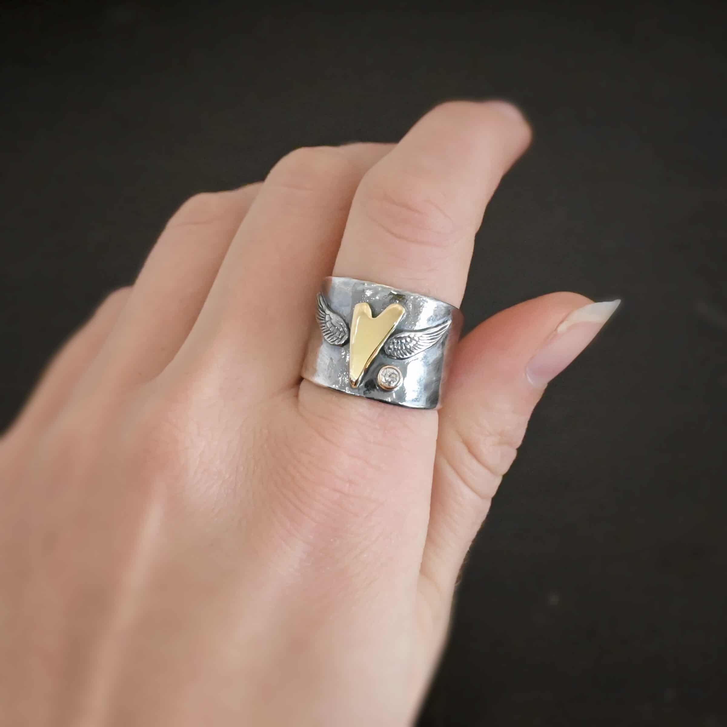 Close up of the "Wild Prairie" Ring. Made out of sterling silver, gold, and diamond. There is a gold heart in the center of the ring. It is a tall heart that stretches from almost the top of the ring to the bottom. There are sterling silver angel wings sprouting from each side of the heart. There is a diamond set in gold on the right side of the ring underneath one of the angel wings. Ring is worn by a model on their pointer finger to demonstrate width of the ring.