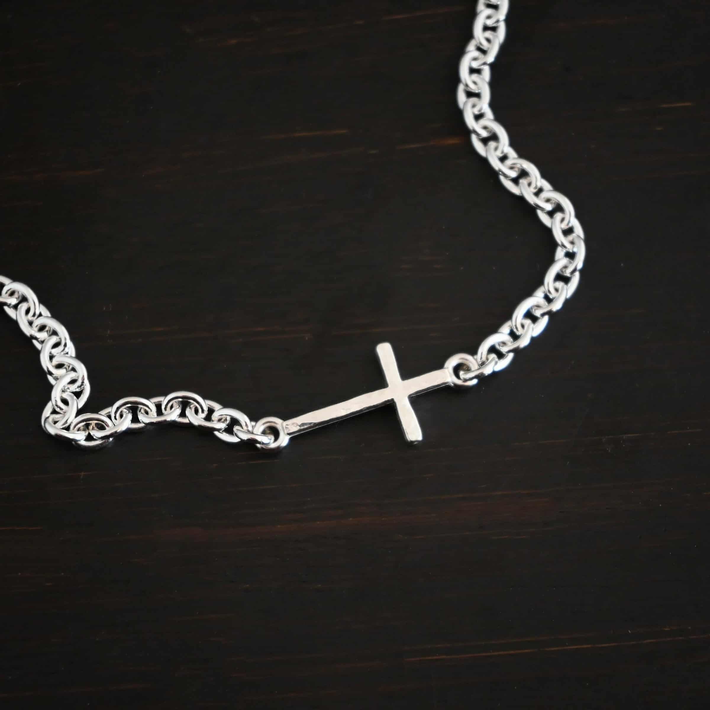 Photo of the "Creator" Bracelet. There is a cross set horizontally with a loop on each end. Each end of the cross is connected to each side of the chain in the center of the bracelet. The bracelet is shiny, and lying against a dark background.