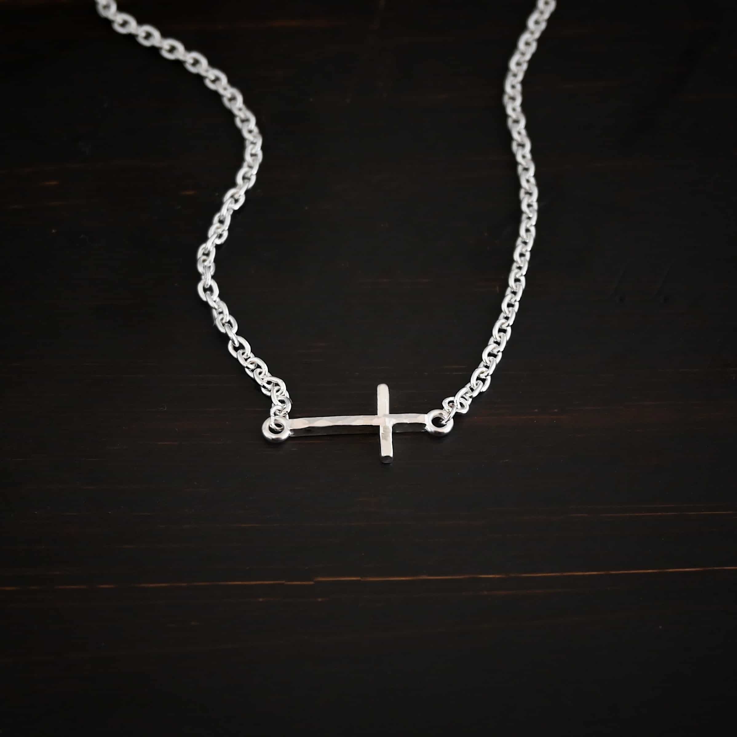 Creator Necklace shiny Photo of the "Creator" Necklace. Made out of sterling silver. There is a small, but thick hammered silver cross hung vertically and attached on both ends to the chain of the necklace. Each end of the cross has a loop on it to attach each end to the chain. Necklace is shiny and lying against a dark background.