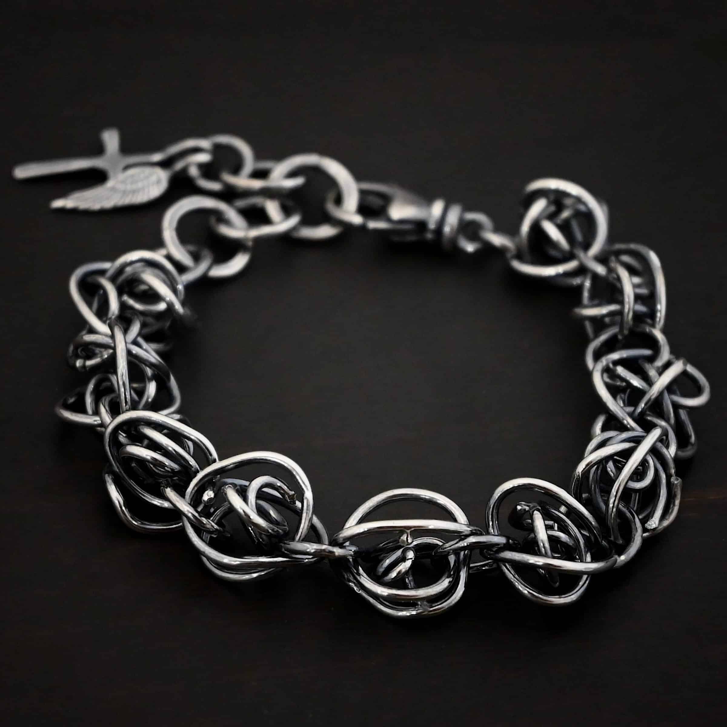 Photo of the "Basket Case" Bracelet. Made out of sterling silver. There are ten, medium sized balls of silver that are all connected closely by jump rings. Each ball is a jumble of sterling silver that wraps around itself multiple times, so it looks crazy and tangled. All of the balls are so close together, that it looks like a continuous wrap. Bracelet has a lobster claw clasp, and a cross and angel wing charm on the back. Bracelet is resting against a dark background.
