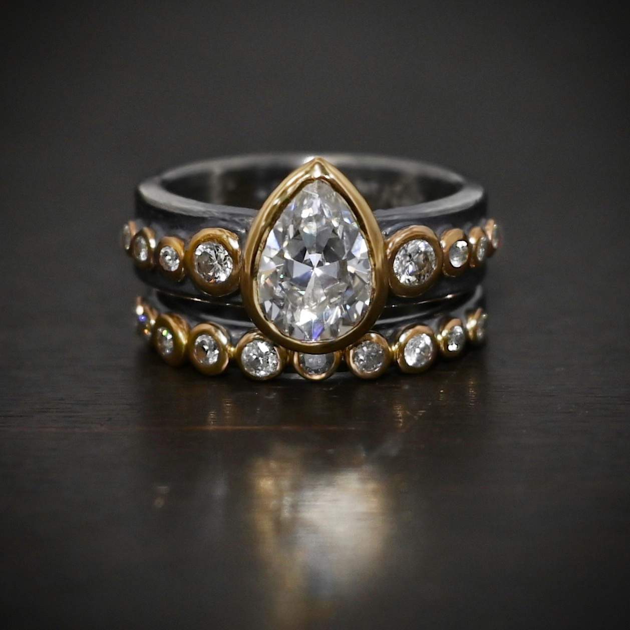 Until Forever Ring Photo of the "Until Forever" Ring. Made out of sterling silver and moissanites. The center moissanite is a pear shape. There are 17 other moissanites that go straight around the top and bottom band. Eight of them are on the top band, four on each side of the pear moissanite. Bands are sitting against a dark background.