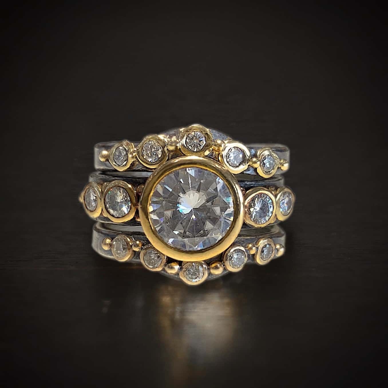 Until Forever Engagement Ring Photo of the "Until Forever" Ring. Made out of sterling silver, gold, and moissanites. There are three separate bands. The top one is curved in the middle and has five moissanites going across it that are set in gold. Each moissanite has a gold ball separating it from the next moissanite. The center band has five moissanites as well. The center is the largest moissanite, and there are two moissanites on the left side and two on the right that are smaller. There is gold ball detail scattered around this band. The last band is also curved in the middle to fit the bottom of the moissanite in the second band. This band has five moissanites, again, separated by gold ball detail. Ring is sitting against a dark background.