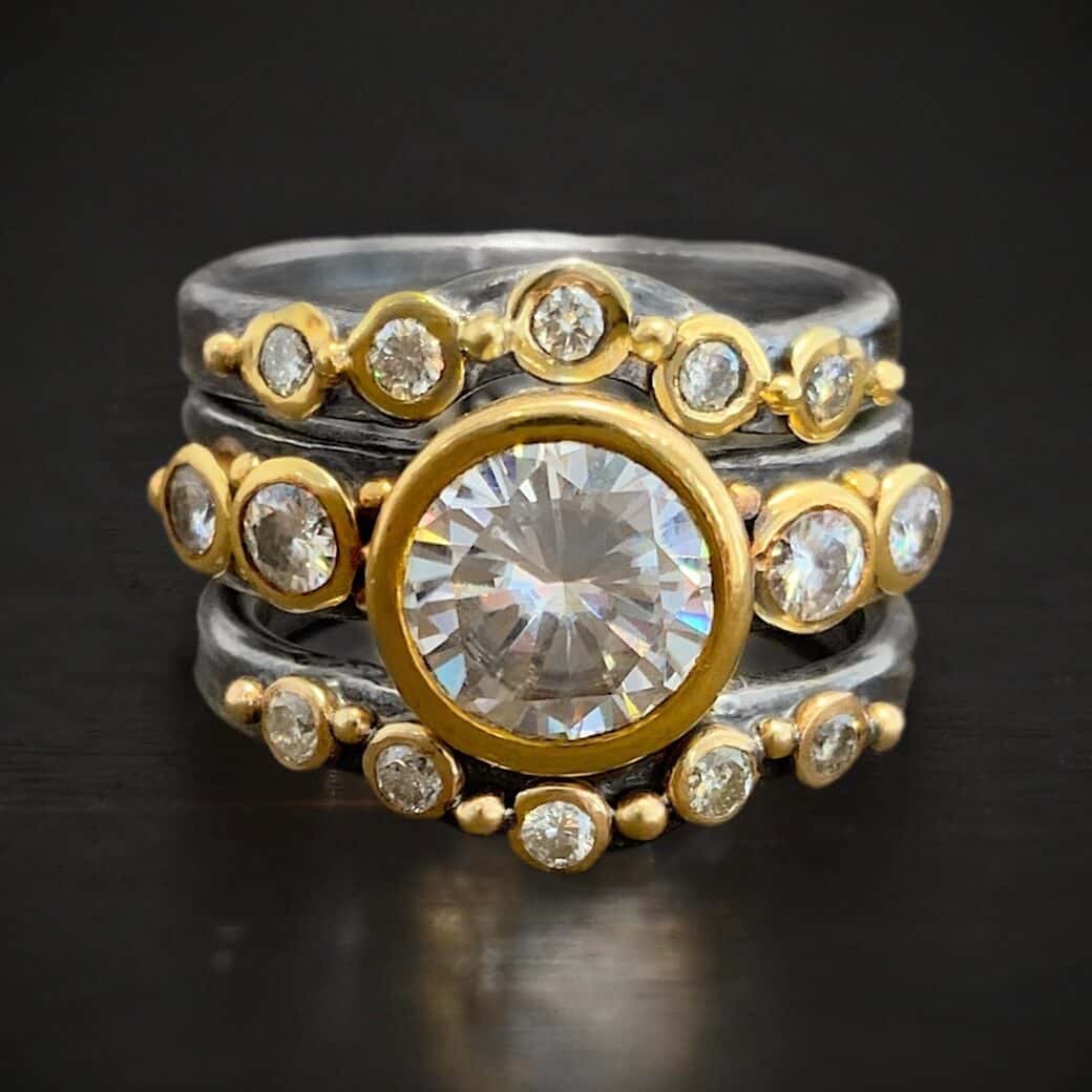 Until Forever Engagement Ring Photo of the "Until Forever" Ring. Made out of sterling silver, gold, and moissanites. There are three separate bands. The top one is curved in the middle and has five moissanites going across it that are set in gold. Each moissanite has a gold ball separating it from the next moissanite. The center band has five moissanites as well. The center is the largest moissanite, and there are two moissanites on the left side and two on the right that are smaller. There is gold ball detail scattered around this band. The last band is also curved in the middle to fit the bottom of the moissanite in the second band. This band has five moissanites, again, separated by gold ball detail. Ring is sitting against a dark background.