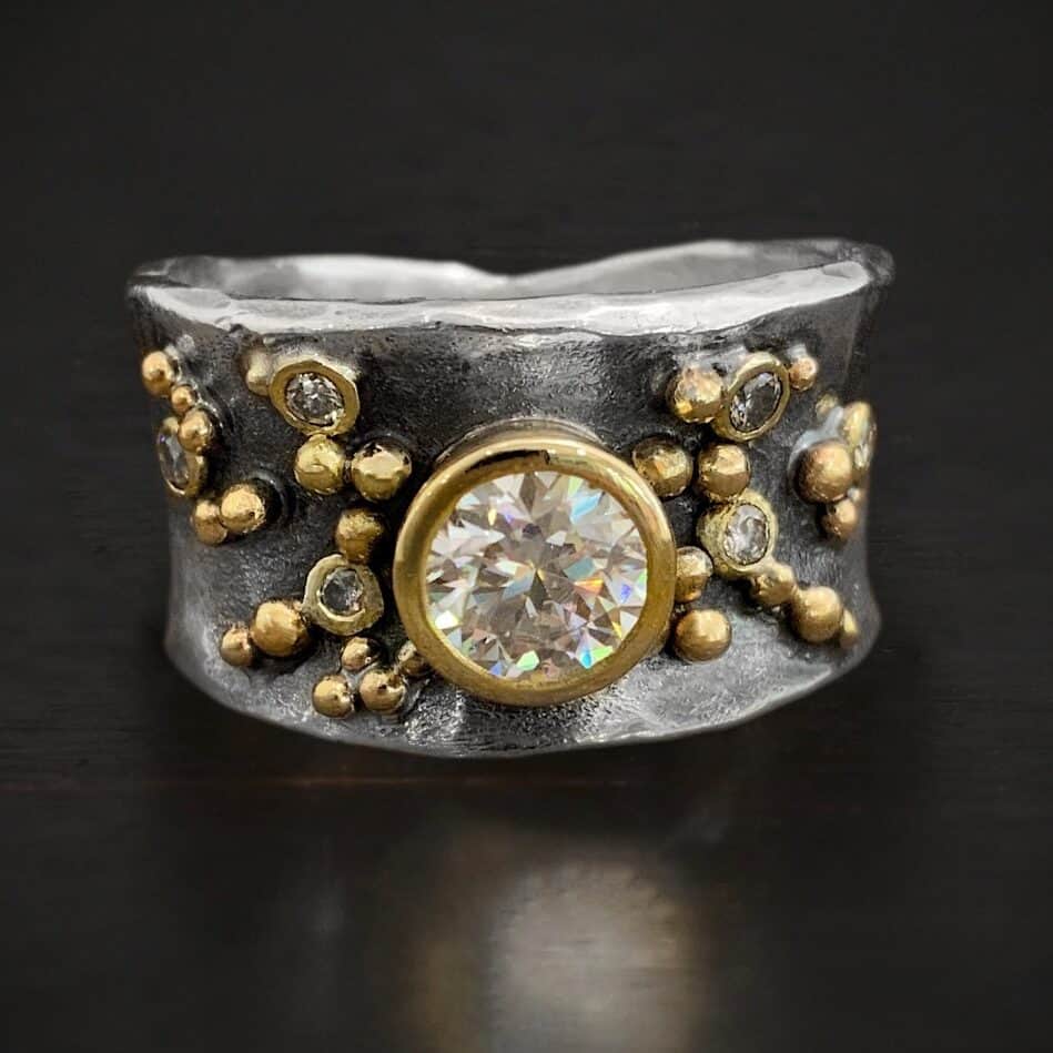 Sunshine & Whiskey Engagement Ring Photo of the "Sunshine & Whiskey" Ring. Made out of sterling silver, gold, and moissanite. The ring has a thick, shiny, silver band that curves slightly inward on the top and bottom. There is a large moissanite in the center of the ring that is set in gold. There are five other moissanites scattered around the ring band. There is a lot of gold ball detail scattered around the ring band as well. Ring is sitting against a dark background.