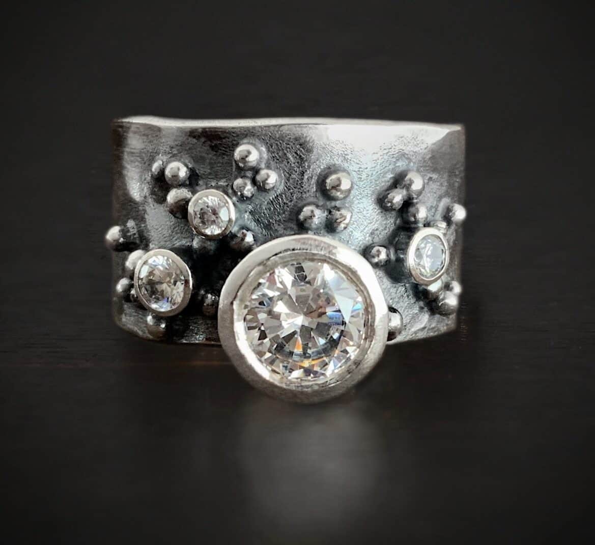 Photo of the Kali Ring. Made out of sterling silver and moissanites. The ring band is thick silver. There are two small moissanites on the right side and one on the left side of the band. There is a large moissanite set near the bottom of the band. The top of the moissanite is soldered to the bottom of the band, so the moissanite slightly sticks off of the bottom of the band. There are balls of silver scattered all around the ring band.