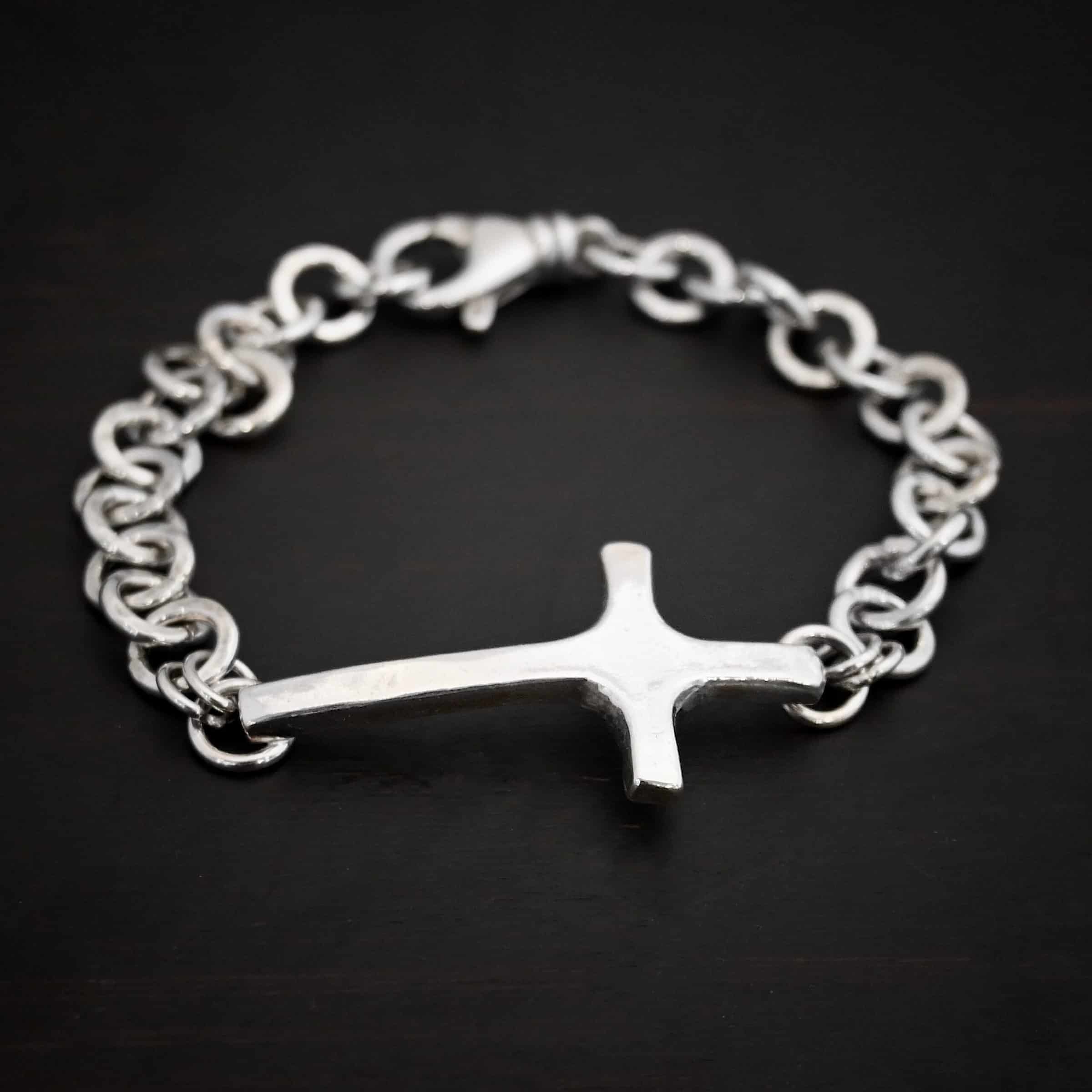 Photo of the "King Of Kings" Bracelet. Made out of sterling silver. There is a thick, medium sized sterling silver cross in the center of the bracelet. The top and bottom of the cross, on the back, have soldered loops. There are two jump rings for each loop, and attached to each jump ring is the thick sterling silver chain of the bracelet. The chain is shiny, thick hammered links. The cross is curved slightly on the bottom to fit around the wrist. Bracelet has a lobster claw clasp. Bracelet is resting against a dark background.