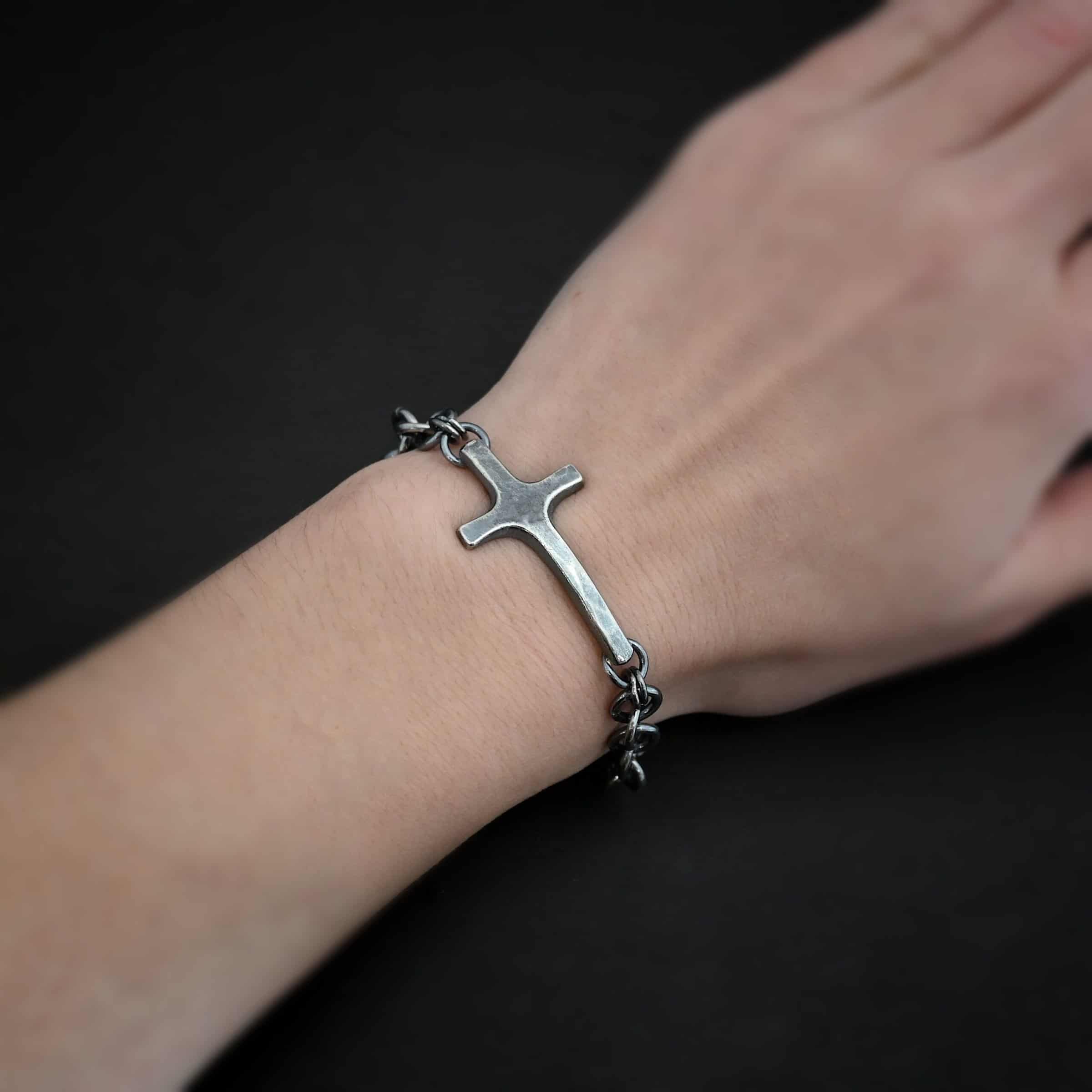 Photo of the "King Of Kings" Bracelet. Made out of sterling silver. There is a thick, medium sized sterling silver cross in the center of the bracelet. The top and bottom of the cross, on the back, have soldered loops. There are two jump rings for each loop, and attached to each jump ring is the thick sterling silver chain of the bracelet. The chain is oxidized, thick hammered links. The cross is curved slightly on the bottom to fit around the wrist. Bracelet has a lobster claw clasp. Bracelet is worn by a model.
