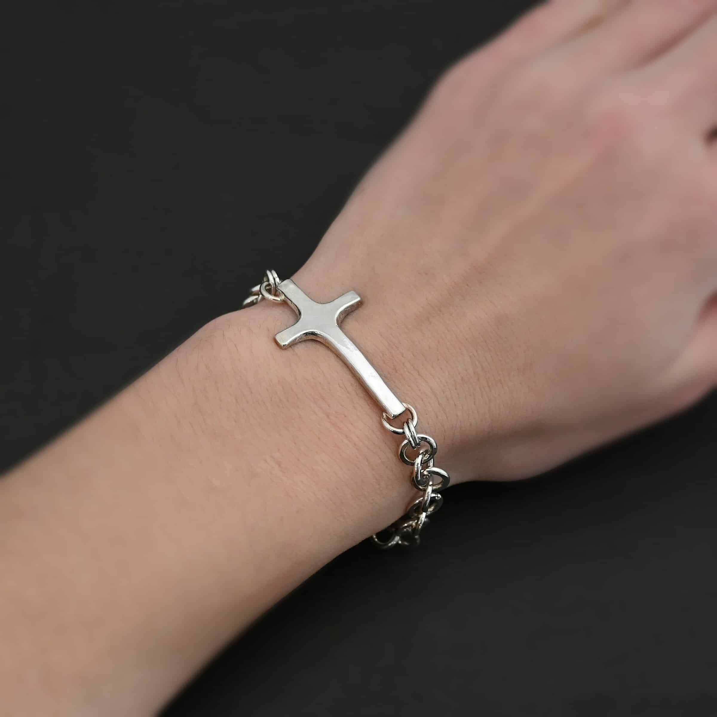 Photo of the "King Of Kings" Bracelet. Made out of sterling silver. There is a thick, medium sized sterling silver cross in the center of the bracelet. The top and bottom of the cross, on the back, have soldered loops. There are two jump rings for each loop, and attached to each jump ring is the thick sterling silver chain of the bracelet. The chain is shiny, thick hammered links. The cross is curved slightly on the bottom to fit around the wrist. Bracelet has a lobster claw clasp. Bracelet is worn by a model.