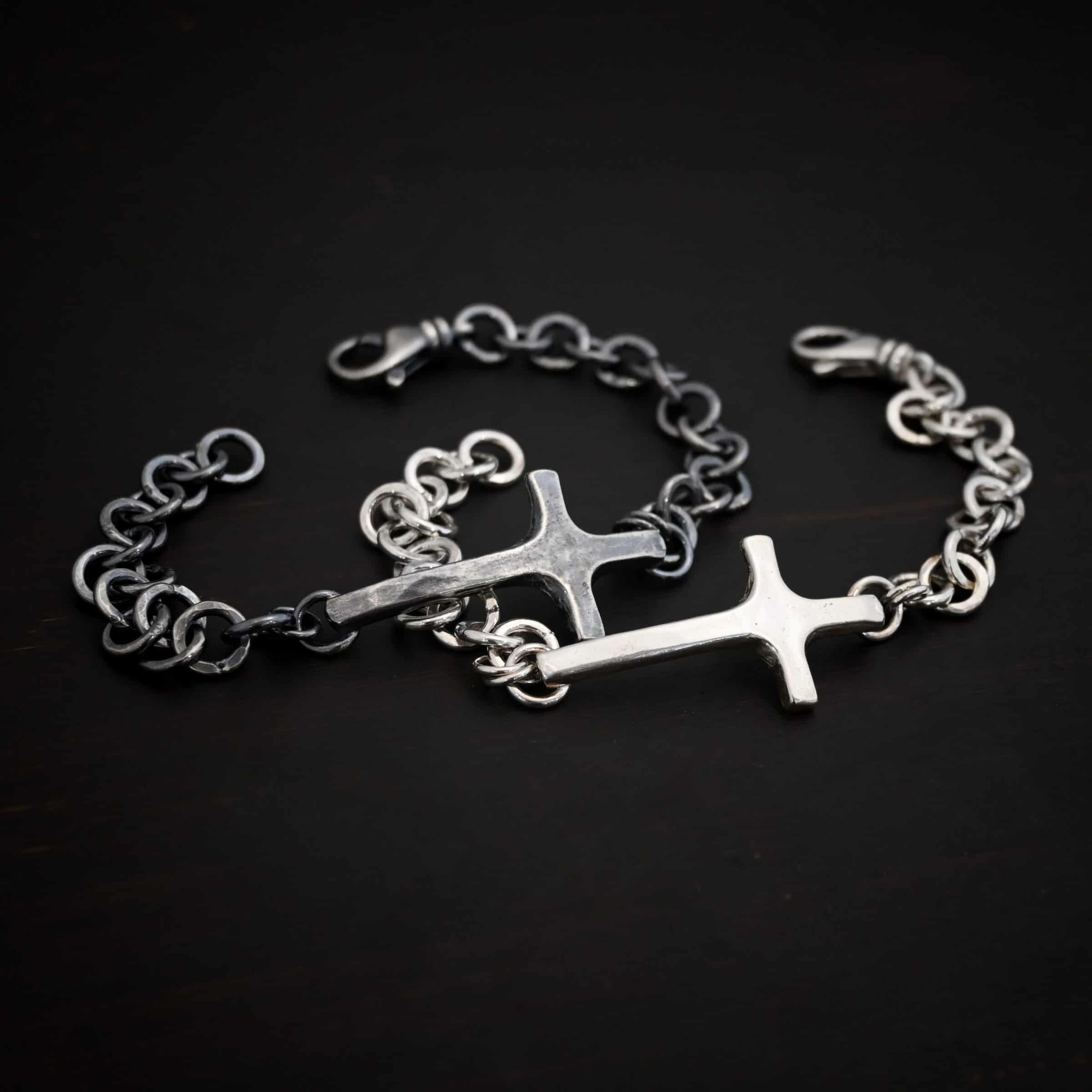 Photo of the "King Of Kings" Bracelet. Made out of sterling silver. There is a thick, medium sized sterling silver cross in the center of the bracelet. The top and bottom of the cross, on the back, have soldered loops. There are two jump rings for each loop, and attached to each jump ring is the thick sterling silver chain of the bracelet. The chain is thick hammered links. The cross is curved slightly on the bottom to fit around the wrist. Bracelet has a lobster claw clasp. Bracelets are lying against a dark background and are shown as both shiny and oxidized.