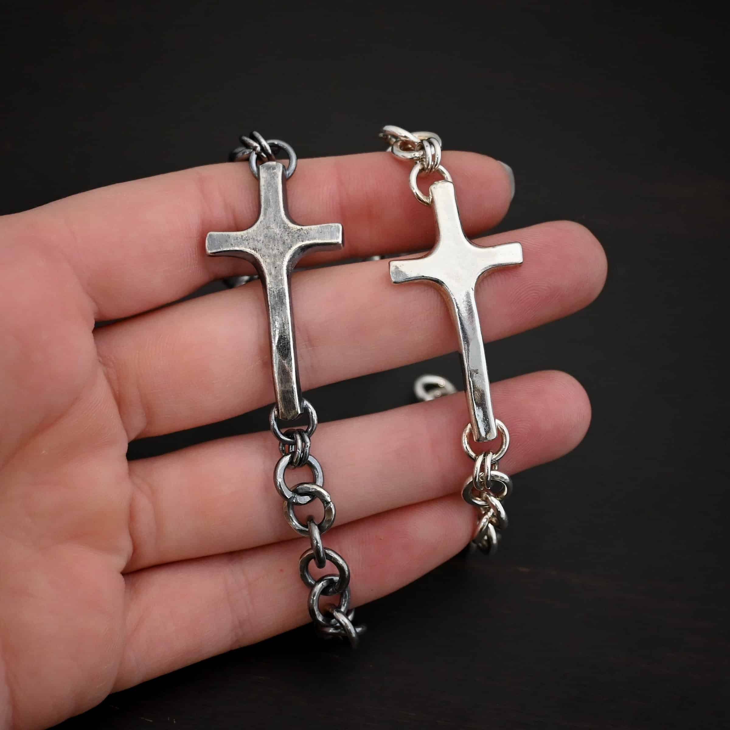 Photo of the "King Of Kings" Bracelet. Made out of sterling silver. There is a thick, medium sized sterling silver cross in the center of the bracelet. The top and bottom of the cross, on the back, have soldered loops. There are two jump rings for each loop, and attached to each jump ring is the thick sterling silver chain of the bracelet. The chain is thick hammered links. The cross is curved slightly on the bottom to fit around the wrist. Bracelet has a lobster claw clasp. Bracelets are lying against the fingertips of a hand and are shown as both shiny and oxidized.