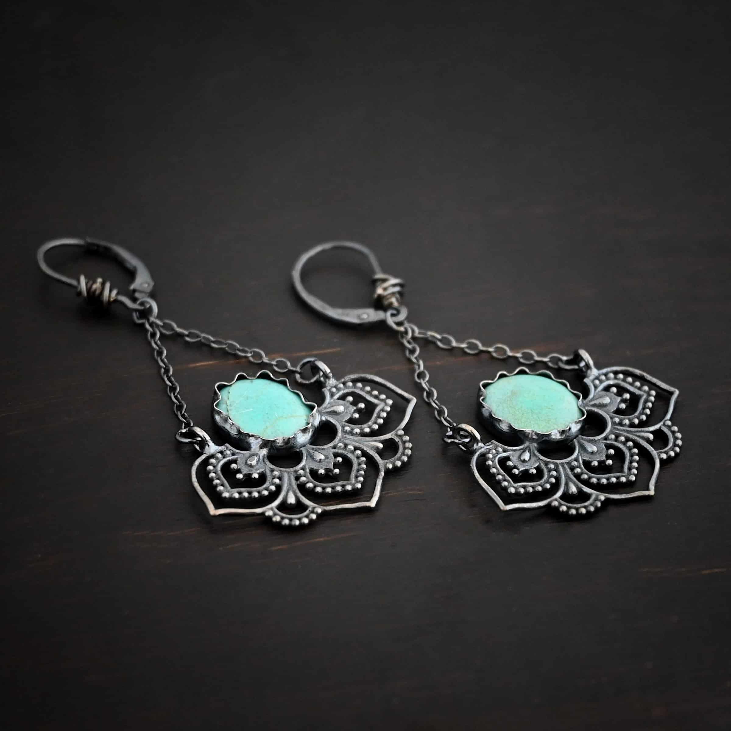Photo of the "Hotel Congress" Earrings. Made out of sterling silver and turquoise. Earrings have leverback ear wires with a jump ring attached to the bottom of them. There are two small strands of silver that are attached to the jump ring. The strands go opposite ways to create a triangle shape. Attached to each end of the strands of silver is a half flower shaped pendant. The pendant is made out of sterling silver, and has three large leaves and two semicircles in between the three leaves. There is an oval turquoise that is attached on the bottom to all three leaves, and the turquoise sits in the center of all three of them. Earrings are lying against a dark background.