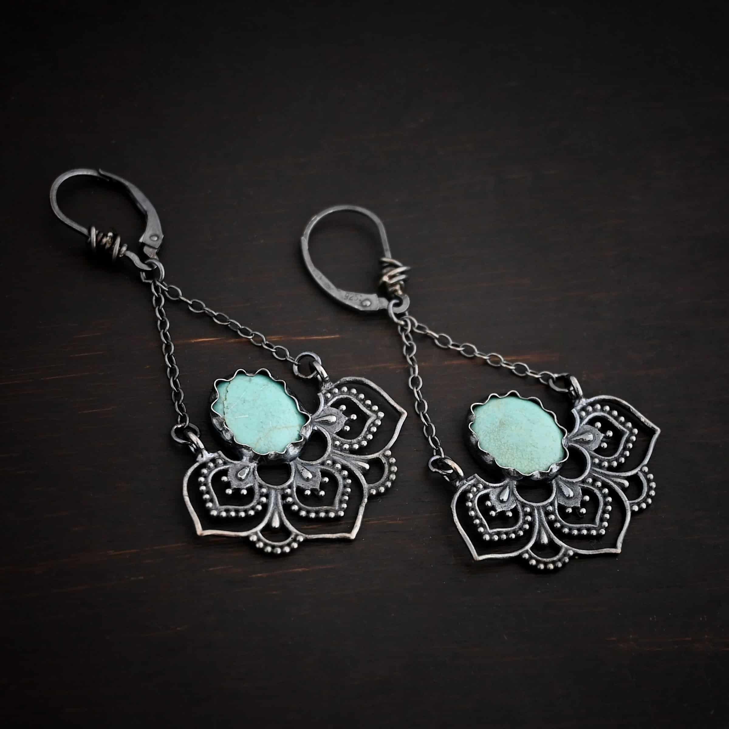 Photo of the "Hotel Congress" Earrings. Made out of sterling silver and turquoise. Earrings have leverback ear wires with a jump ring attached to the bottom of them. There are two small strands of silver that are attached to the jump ring. The strands go opposite ways to create a triangle shape. Attached to each end of the strands of silver is a half flower shaped pendant. The pendant is made out of sterling silver, and has three large leaves and two semicircles in between the three leaves. There is an oval turquoise that is attached on the bottom to all three leaves, and the turquoise sits in the center of all three of them. Earrings are lying against a dark background.