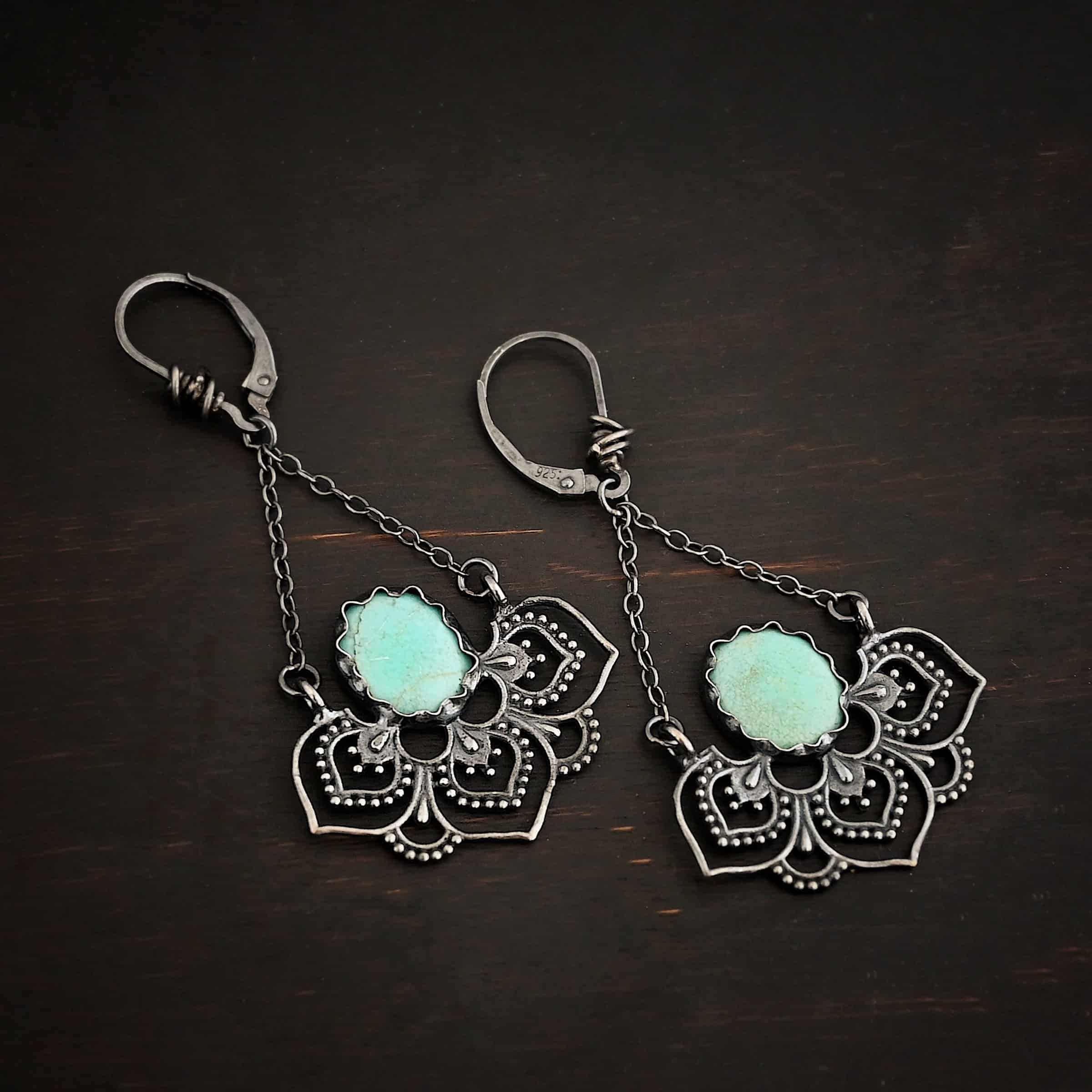 Photo of the "Hotel Congress" Earrings. Made out of sterling silver and turquoise. Earrings have leverback ear wires with a jump ring attached to the bottom of them. There are two small strands of silver that are attached to the jump ring. The strands go opposite ways to create a triangle shape. Attached to each end of the strands of silver is a half flower shaped pendant. The pendant is made out of sterling silver, and has three large leaves and two semicircles in between the three leaves. There is an oval turquoise that is attached on the bottom to all three leaves, and the turquoise sits in the center of all three of them. Earrings are lying against a dark background.