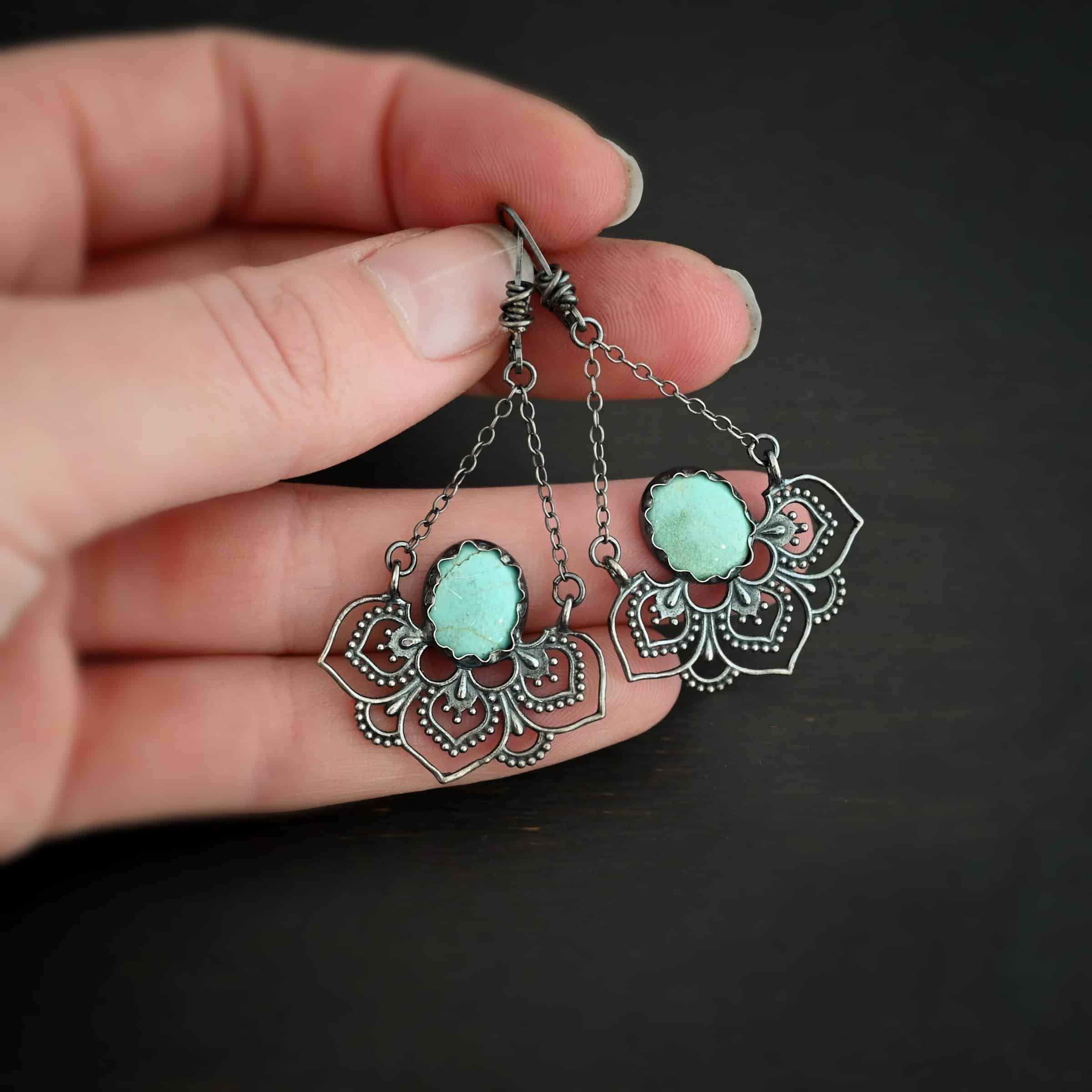 Photo of the "Hotel Congress" Earrings. Made out of sterling silver and turquoise. Earrings have leverback ear wires with a jump ring attached to the bottom of them. There are two small strands of silver that are attached to the jump ring. The strands go opposite ways to create a triangle shape. Attached to each end of the strands of silver is a half flower shaped pendant. The pendant is made out of sterling silver, and has three large leaves and two semicircles in between the three leaves. There is an oval turquoise that is attached on the bottom to all three leaves, and the turquoise sits in the center of all three of them. Earrings are lying against a dark background.