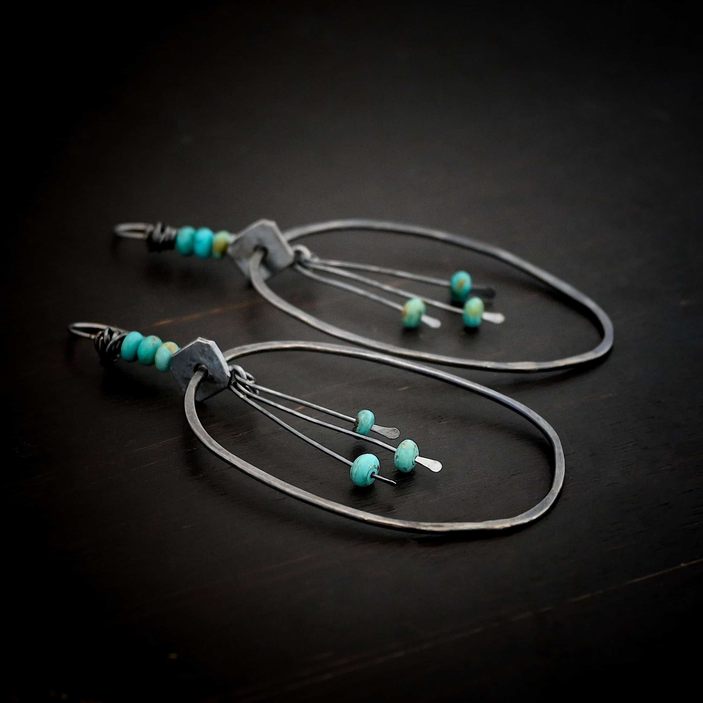 Photo of the "Montana Sky" Earrings. Made out of sterling silver and turquoise. Earrings have french hook ear wires with silver wrapped around the top of them. There are three turquoise beads attached to the top of the ear wires as well. After the turquoise, there is a square, thin piece of silver set sideways with a hole in the center. Going through the hole is a large oval hoop that is hammered flat, but smooth. Hanging from the bottom of the square off of a soldered jump ring, in the center of the oval are three small, thin sticks of silver with loops on the top and hammered paddles on the bottom. Hanging just above the paddles on each stick is a small turquoise bead. Earrings are lying against a dark background.